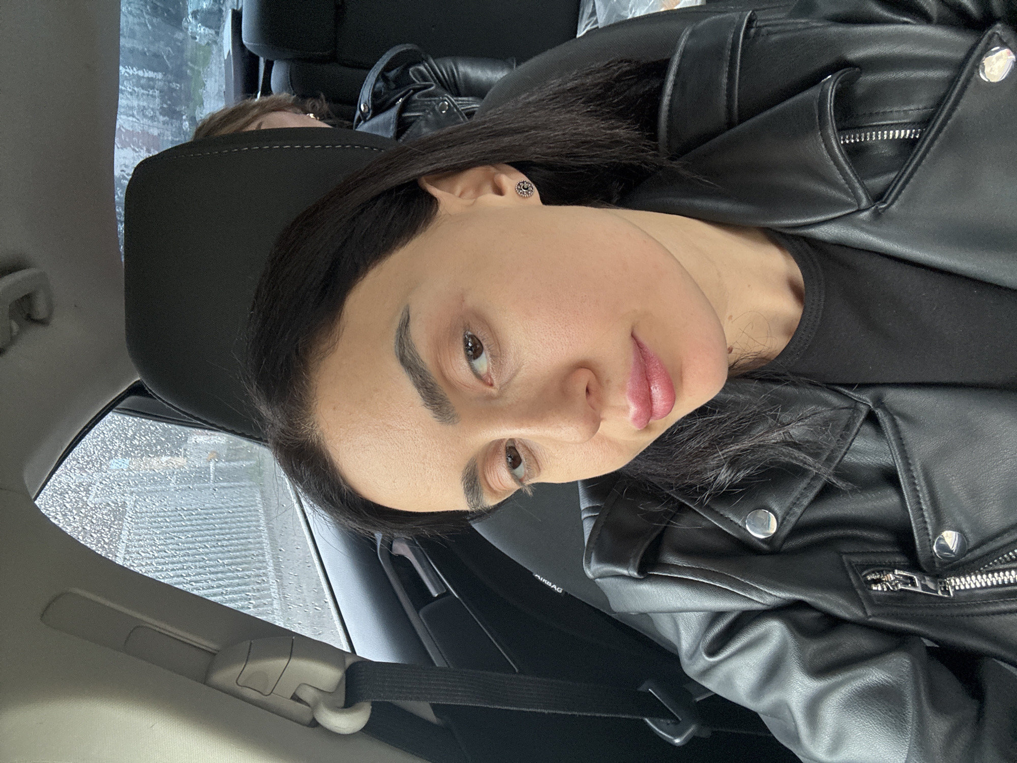 A woman with dark hair and light skin is in the passenger seat of a car. She is wearing a black leather jacket over a black shirt and has a stud earring in her left ear. The seatbelt is fastened across her chest. Rain is visible on the car windows. Another person's head is visible in the background, behind the car seat. The location appears to be in Tbilisi, Georgia based on environmental cues.