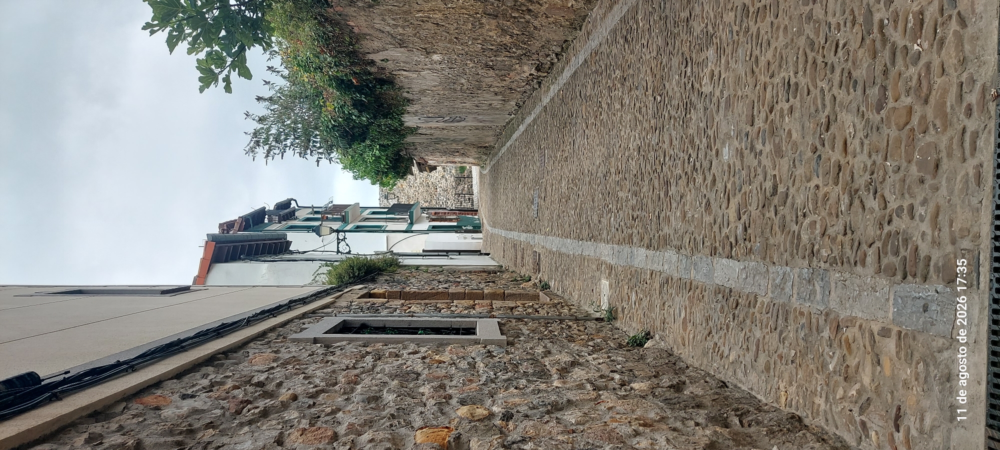 This is a vertical, eye-level, full shot of a cobblestone street in Castro-Urdiales, Spain, on a cloudy day. The street is lined with stone buildings on the left and a stone wall on the right. The buildings on the left are two-story with stone lower walls and white upper walls. The building on the right is a rough-hewn stone wall, topped with lush green foliage that drapes over the edge. A white stripe runs down the center of the cobblestone street. The street curves gently to the right and recedes into the distance, where a metal fence and more stone architecture are visible. The sky is overcast. The timestamp "11 de agosto de 2026 17:35" is overlaid on the bottom left corner of the image. There are no people or vehicles visible in the scene.