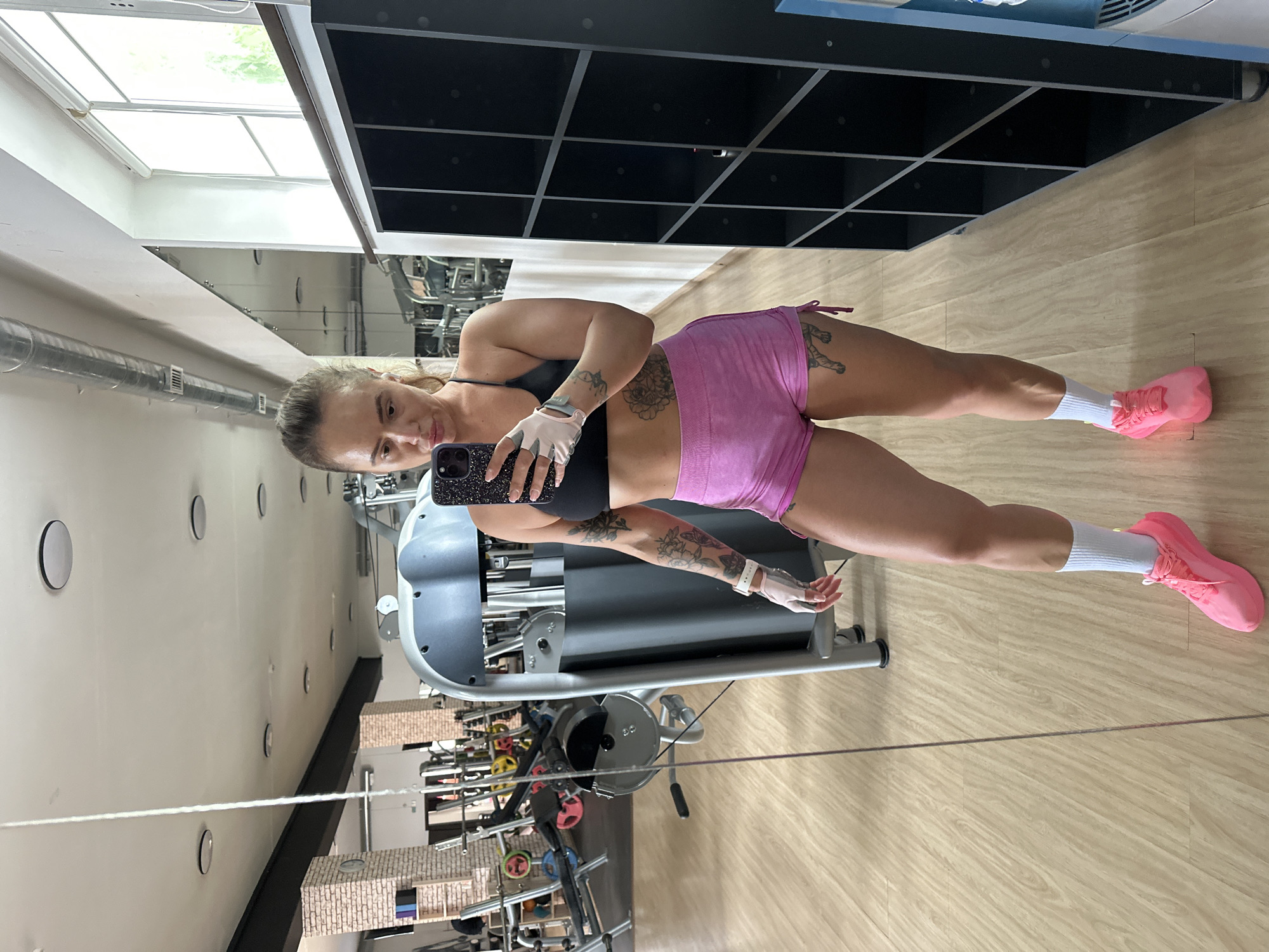 A full-body selfie is captured in a gym in Yuzhnouralsk, Russia. A woman, wearing a black sports bra, pink shorts, white socks, and pink athletic shoes, stands facing a mirror and holding a smartphone. She has blonde hair pulled back in a ponytail and is wearing fingerless gloves. Tattoos are visible on her arms and thighs. The gym environment includes exercise equipment, a large mirror reflecting the room, and a black locker unit. The floor is made of light-colored wood. Natural light streams in from windows on the right side of the image.