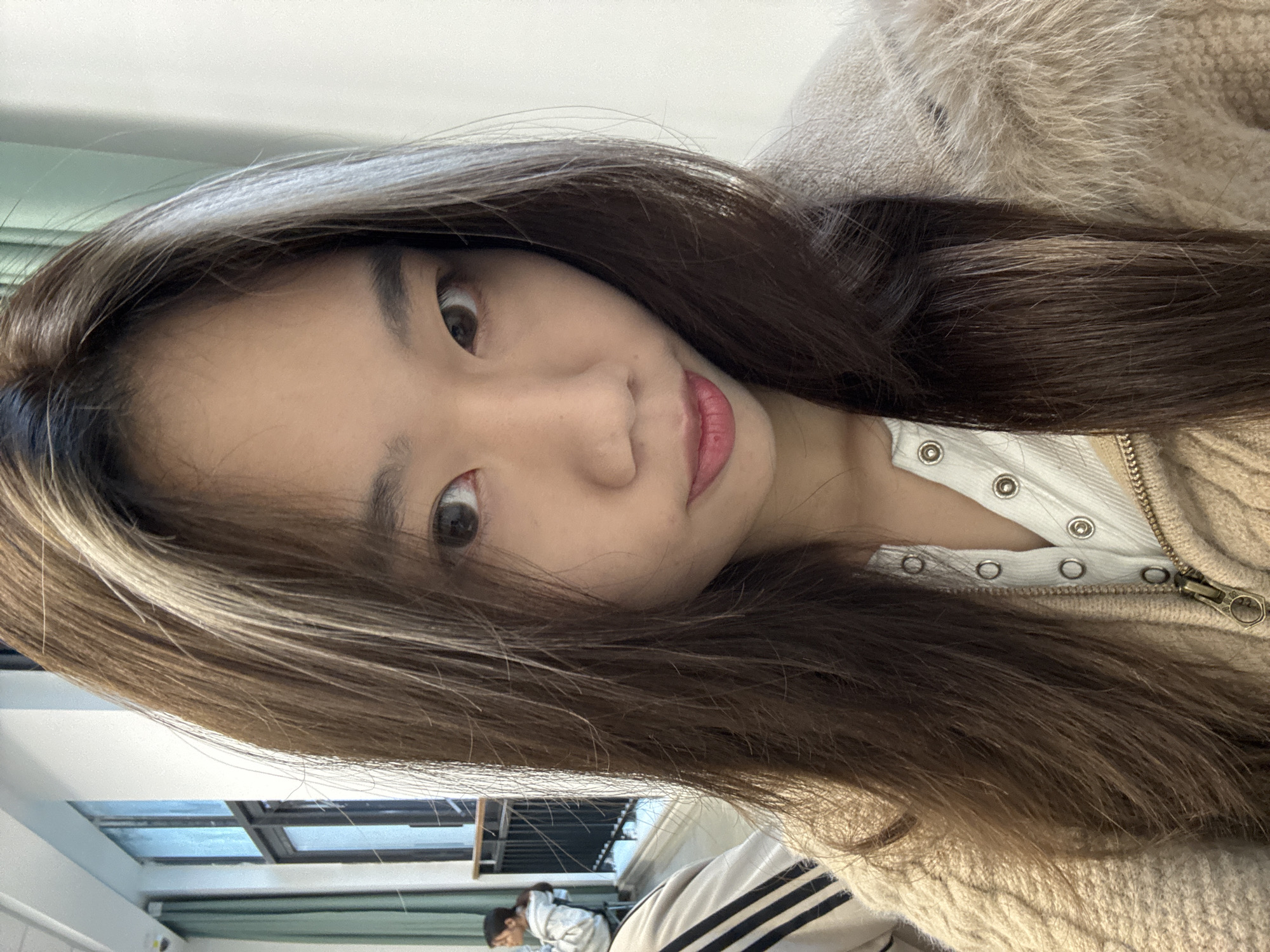 The image presents a close-up, chest-up selfie of a young East Asian female looking directly into the camera with a slight smirk. She has long, dark brown hair with lighter, possibly highlighted, streaks framing her face. She is wearing a light-colored, possibly white, ribbed top with metallic snap buttons, beneath a beige cable-knit jacket with a brass-colored zipper. A light brown faux fur trim or collar of the jacket is visible on her right shoulder.
In the blurred background to the left, another female with dark hair, wearing a light grey hooded sweatshirt, is partially visible from the back, seated. A segment of a light-colored garment with three dark horizontal stripes is present in the lower-left foreground, likely belonging to a third person or the photographer. The mid-ground features a multi-pane window structure with green curtains or blinds, a black metal railing, and a wooden handrail, indicating an indoor balcony or corridor. A white wall, a white ceiling with a rectangular light fixture, and a small, white dome-shaped security camera are also visible in the upper-left. The background on the right side of the main subject is a plain white wall. The scene is an interior public or institutional space, located in Nan'an District, China.