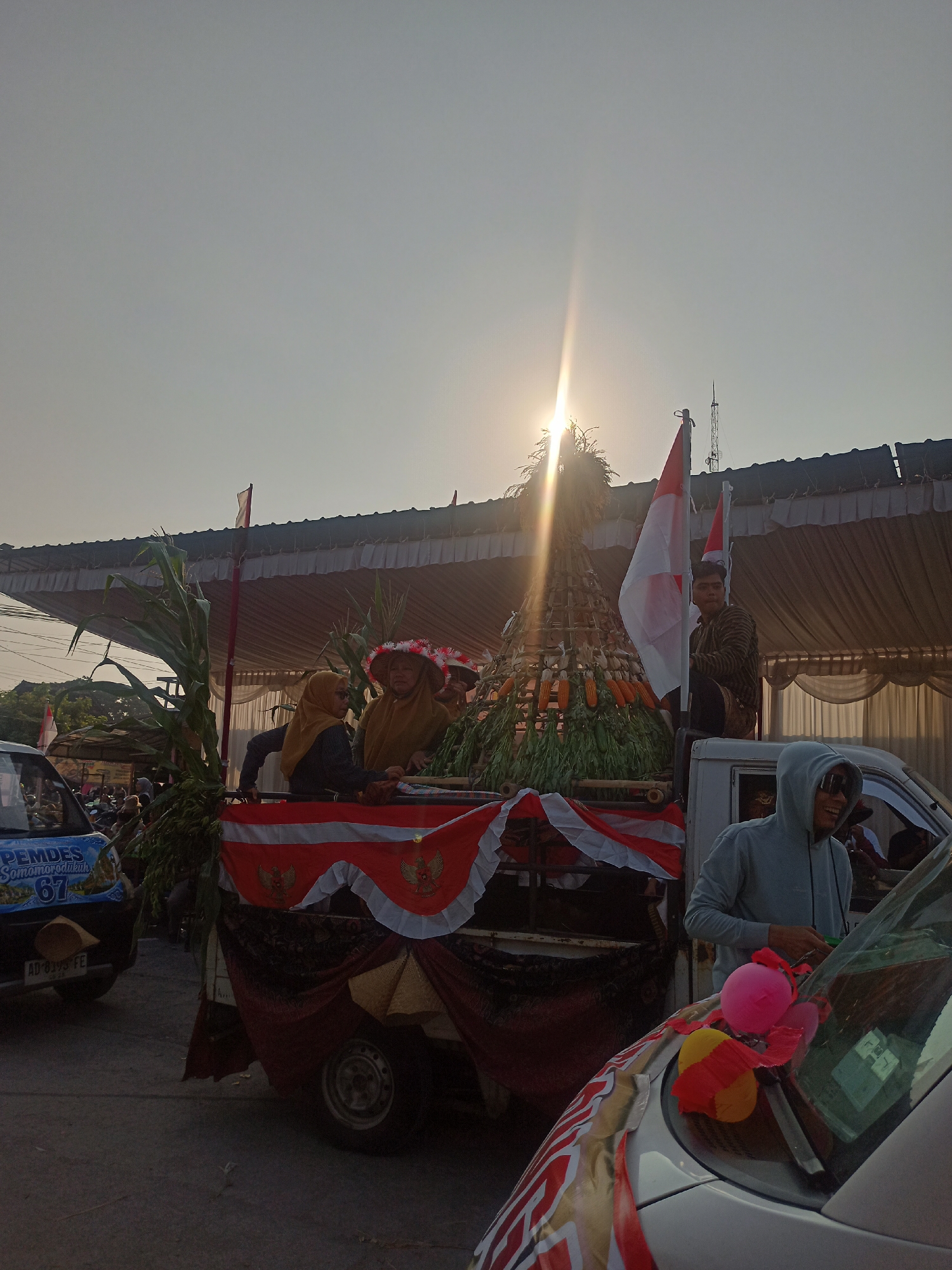 The image captures a scene in Ngrompongan, Indonesia, likely during a celebratory event. A decorated flatbed truck is the central focus, adorned with red and white Indonesian flags and a conical structure made of bamboo, filled with corn and other agricultural produce. Several individuals are visible on the truck, some wearing traditional attire and hats.
In the foreground, a white car is partially visible, with balloons attached to its hood, suggesting a festive atmosphere. The sunlight is strong, casting a lens flare and illuminating the sky, which is clear with a slight haze. In the background, a building with a large awning and some trees can be seen. Another vehicle, a blue van with "PEMDES Somoroodukuh 67" written on it, is parked to the left of the truck. The overall impression is one of a community gathering, possibly a parade or festival, showcasing local produce and national pride.