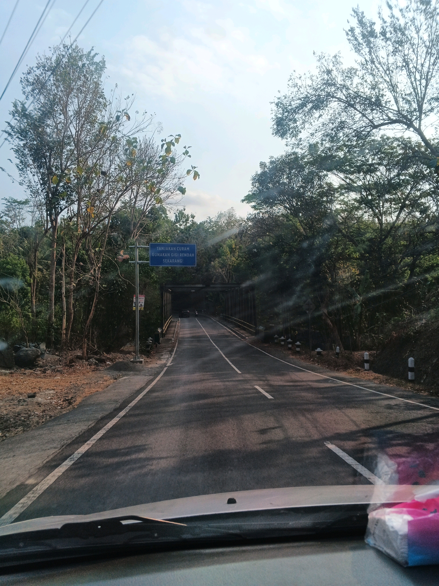 The image shows a road curving through a forested area in Minggir, Indonesia. A blue sign with white text is visible on the left side of the road, reading "TANJAKAN CURAM GUNAKAN GIGI RENDAH SEKARANG!" which translates to "STEEP CLIMB USE LOW GEAR NOW!". A bridge spans the road ahead, with trees lining both sides of the road and a clear sky overhead. The perspective suggests the photo was taken from inside a vehicle, as the dashboard and windshield wipers are visible in the foreground.
