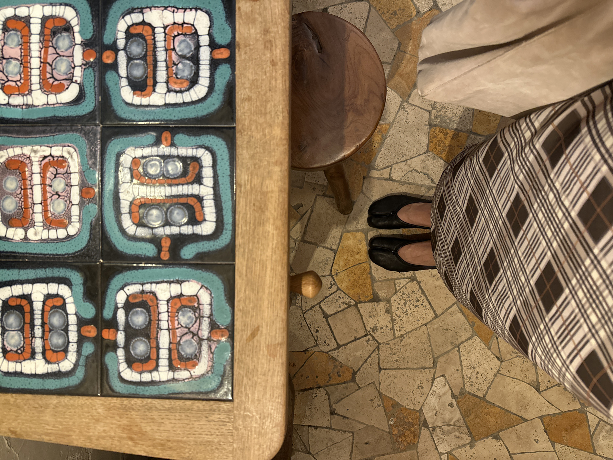 The image shows a top-down view of a tiled table with decorative, abstract geometric patterns in turquoise, orange, white, and black. Next to the table is a small, round wooden stool with three legs. To the right of the stool, a person's legs are visible, wearing black split-toe tabi-style shoes and a brown and white plaid skirt. A light-colored fabric, possibly a coat or blanket, is draped over the person. The floor is covered in irregular, earth-toned mosaic tiles. The scene appears to be indoors, possibly in a cafe or home, with natural light casting shadows. The overall impression is one of a quiet, domestic or commercial setting. No text is visible in the image. The context of Moscow, Russia, is provided but not visually evident in the image itself.
