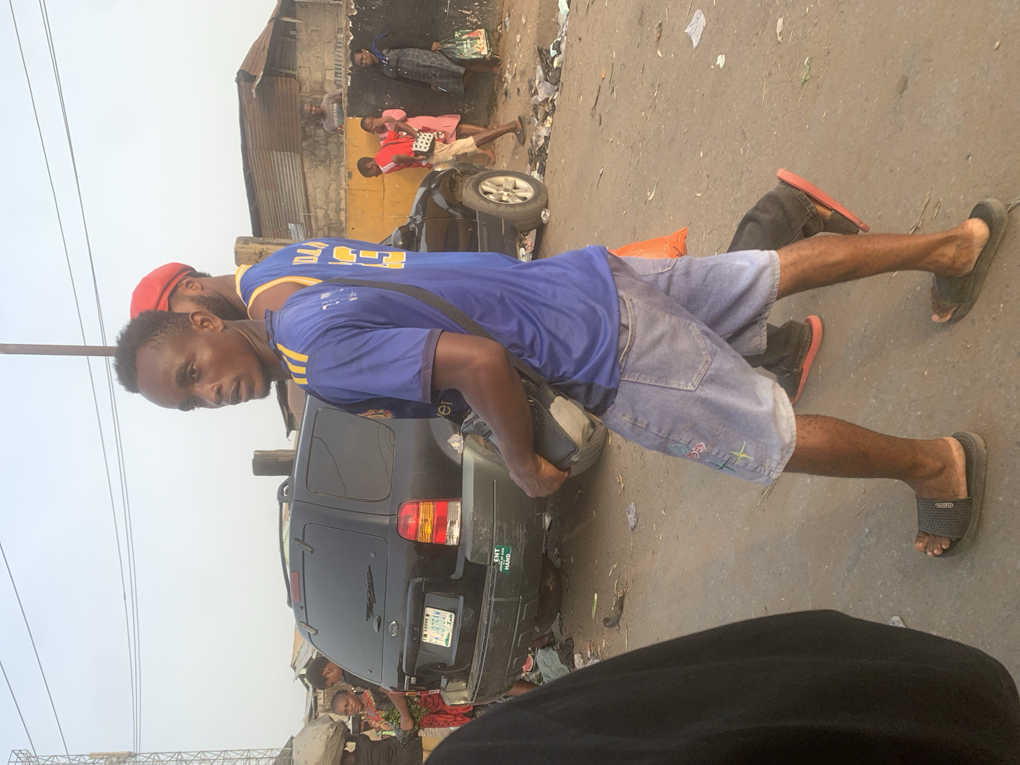 This media file captures a busy street scene, likely in an urban area, viewed from a low angle. The primary subject is a young man with dark skin, positioned in the foreground and looking towards the left. He wears a blue sleeveless jersey with yellow accents (resembling a basketball jersey with "D.L." and a number partially visible), denim shorts, and dark flip-flops. A dark shoulder bag hangs across his body. Another person, partially visible behind him, wears a red cap.
In the midground, a dark-colored SUV, possibly a Ford Escape, is parked with its rear facing the viewer. Its license plate, "LAGOS FKJ 975AM," is clearly visible, indicating the location is Lagos, Nigeria. A sticker on the bumper of the SUV shows partial text including "ENT," "GOOD," and "HARD." Several other individuals are present: one person in a red shirt and light shorts carries a white handbag, and another in a patterned dress stands further back. The ground is a mix of asphalt and dirt, with visible litter.
The background consists of simple, single-story buildings with corrugated iron roofs and concrete walls, suggesting a residential or commercial district. Overhead, utility lines or structures are visible against a bright, overcast sky, indicating it is daytime. The scene portrays an everyday street environment with multiple people and vehicles. The flip-flops worn by the main subject have the brand name "FUOFU" printed on them.