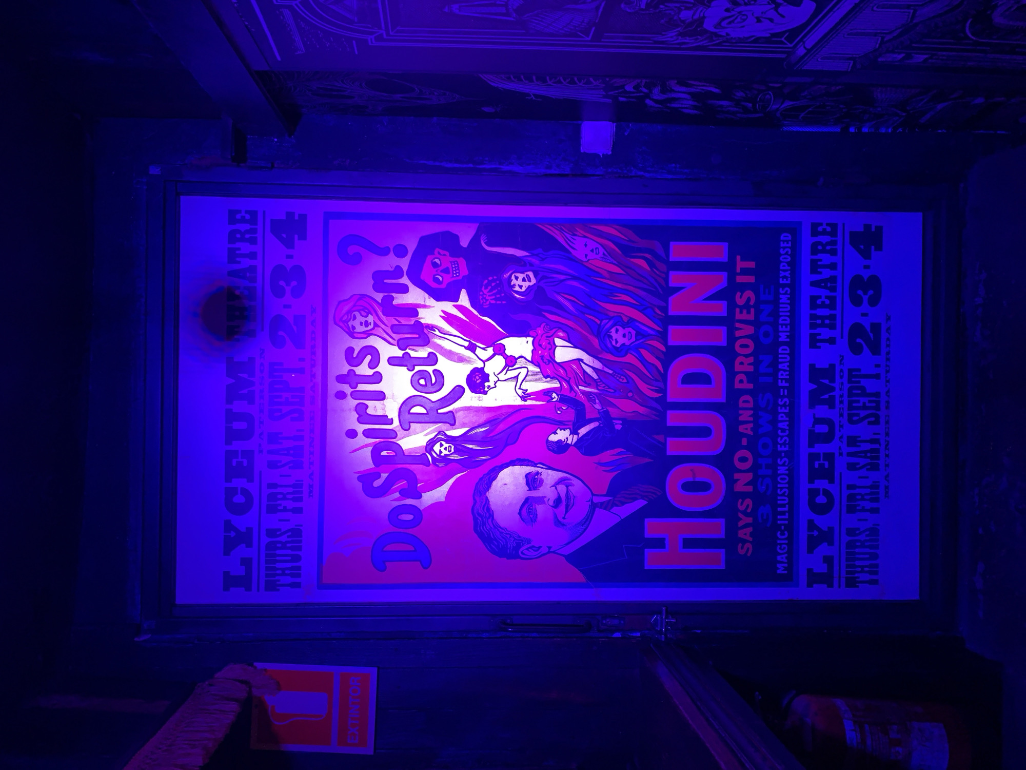 A framed poster advertising Harry Houdini's show, "Do Spirits Return?" at the Paterson Lyceum Theatre, is hung on a dark wall. The poster is lit with blue light. An "EXTINTOR" (fire extinguisher) sign is visible on the lower left, with a fire extinguisher itself partially visible on the lower right. The background is dark and appears to be an interior space with decorative paneling visible at the top. The image was taken indoors under poor lighting conditions. No people are visible in the frame.