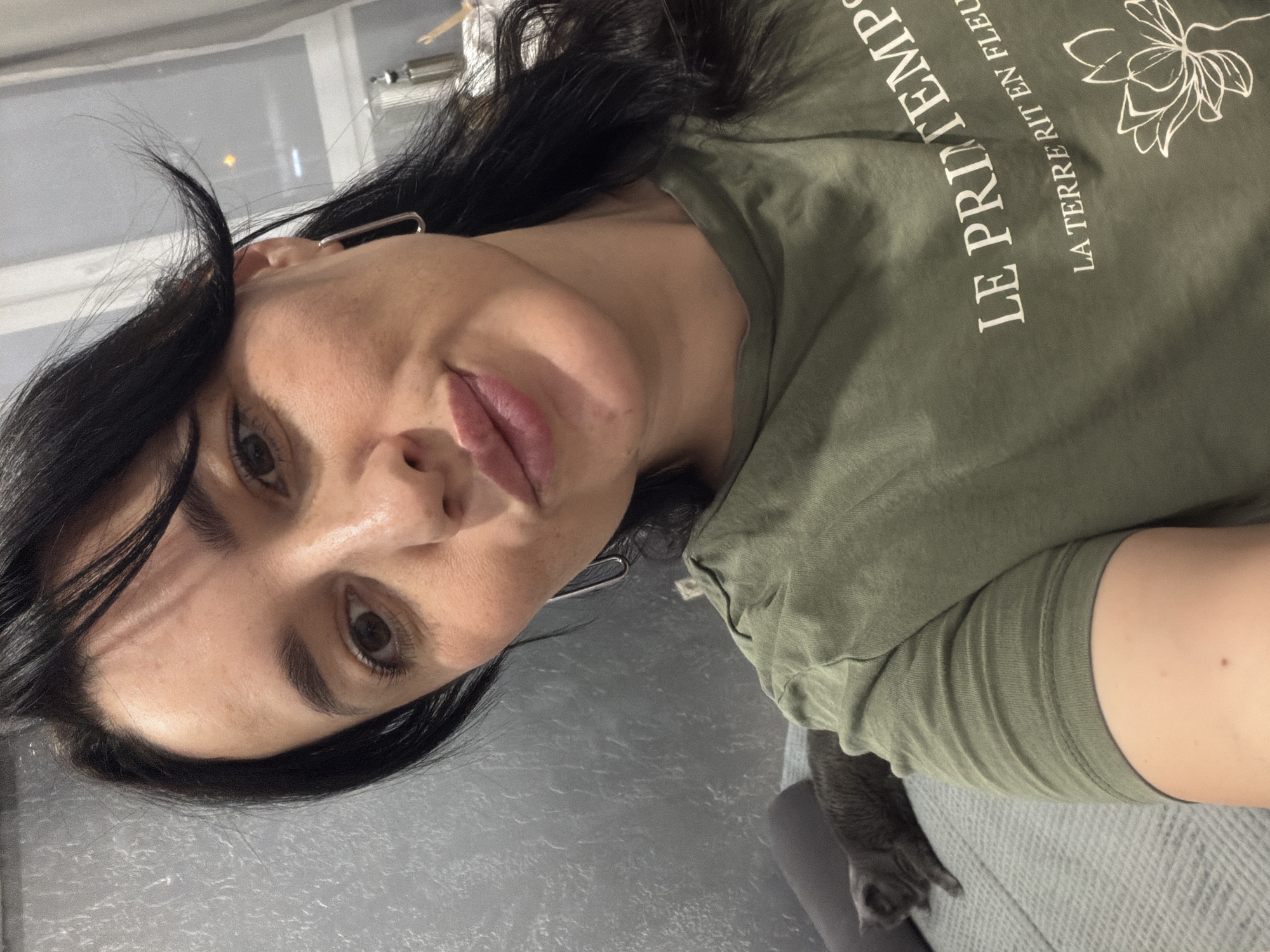 The image is a selfie of a woman with dark hair and brown eyes, wearing a green t-shirt with white text that reads "LE PRINTEMPS LA TERRE RIT EN FLEUR". She is making a kissy face. A grey cat is visible on a grey couch in the background. The setting appears to be an indoor residential environment, possibly an apartment in the city of Korolyov, Russia.