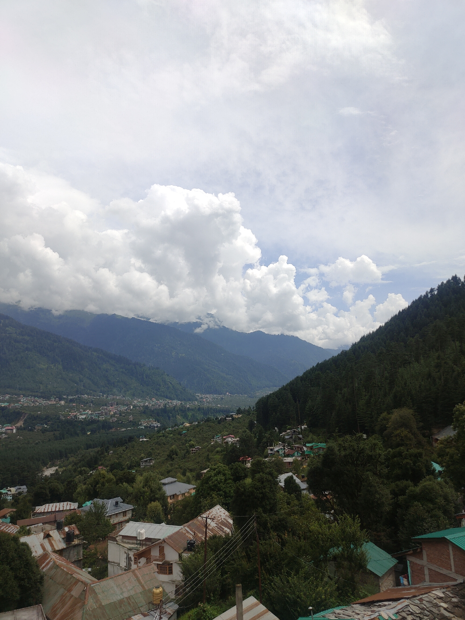 The image depicts a panoramic view of Manali, India, showcasing its characteristic hilly terrain and a densely populated settlement nestled within the valley. The foreground is dominated by a cluster of buildings with varying roof types, including corrugated metal and slate, suggesting a mix of residential and commercial structures. Some buildings appear older with weathered roofs, while others exhibit more modern construction. Trees and vegetation are interspersed among the buildings, creating a harmonious blend of nature and human habitation.
In the mid-ground, the landscape transitions to a sprawling town or city, with numerous buildings extending across the valley floor. The distinct architectural styles and varied roof colors of these structures are visible, hinting at the density and diversity of the urban fabric. Beyond the settlement, imposing mountains rise, their slopes covered with dense coniferous forests. The lower slopes of these mountains appear to be cultivated or cleared in some areas, revealing patches of green.
The background is characterized by the majestic Himalayan mountain range. The peaks are shrouded in a dramatic sky filled with large, fluffy white clouds that cast shadows on the mountainside, creating a sense of depth and scale. The overall atmosphere of the image is one of a tranquil yet vibrant mountain town, bathed in the soft light of an overcast day. The scene conveys the impression of a settled community thriving amidst a breathtaking natural landscape.