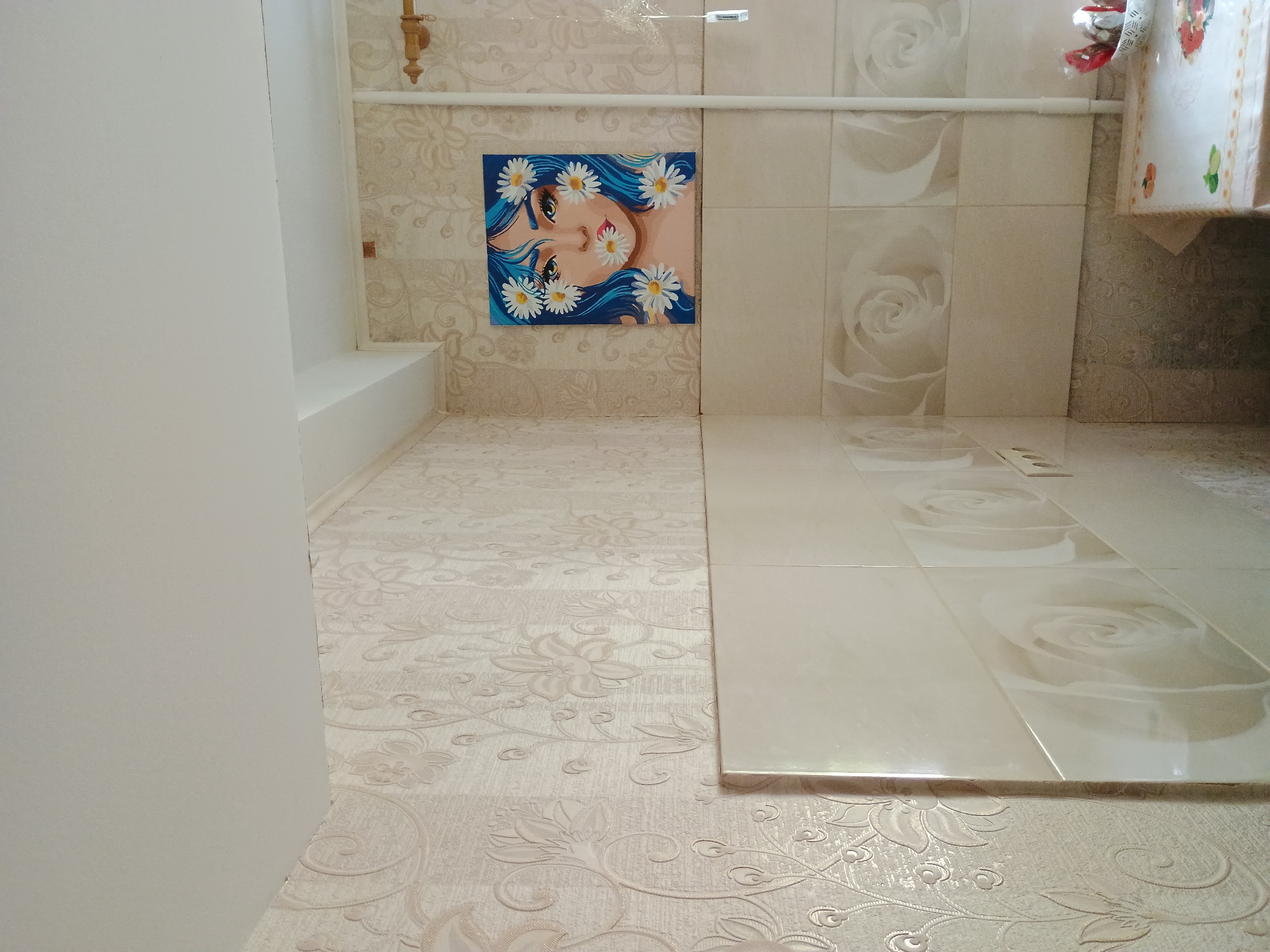 The image shows a corner of a room, likely a kitchen, with floral patterned wallpaper and tiled walls. On the wall, there is a painting of a woman with blue hair and daisies around her face. A radiator pipe runs vertically along the right side of the image. A table covered with a tablecloth is visible in the lower right corner. The scene appears to be in an interior space, and no specific location in Zaporizhzhia, Ukraine can be determined from the visual evidence. There are no people present in the image.