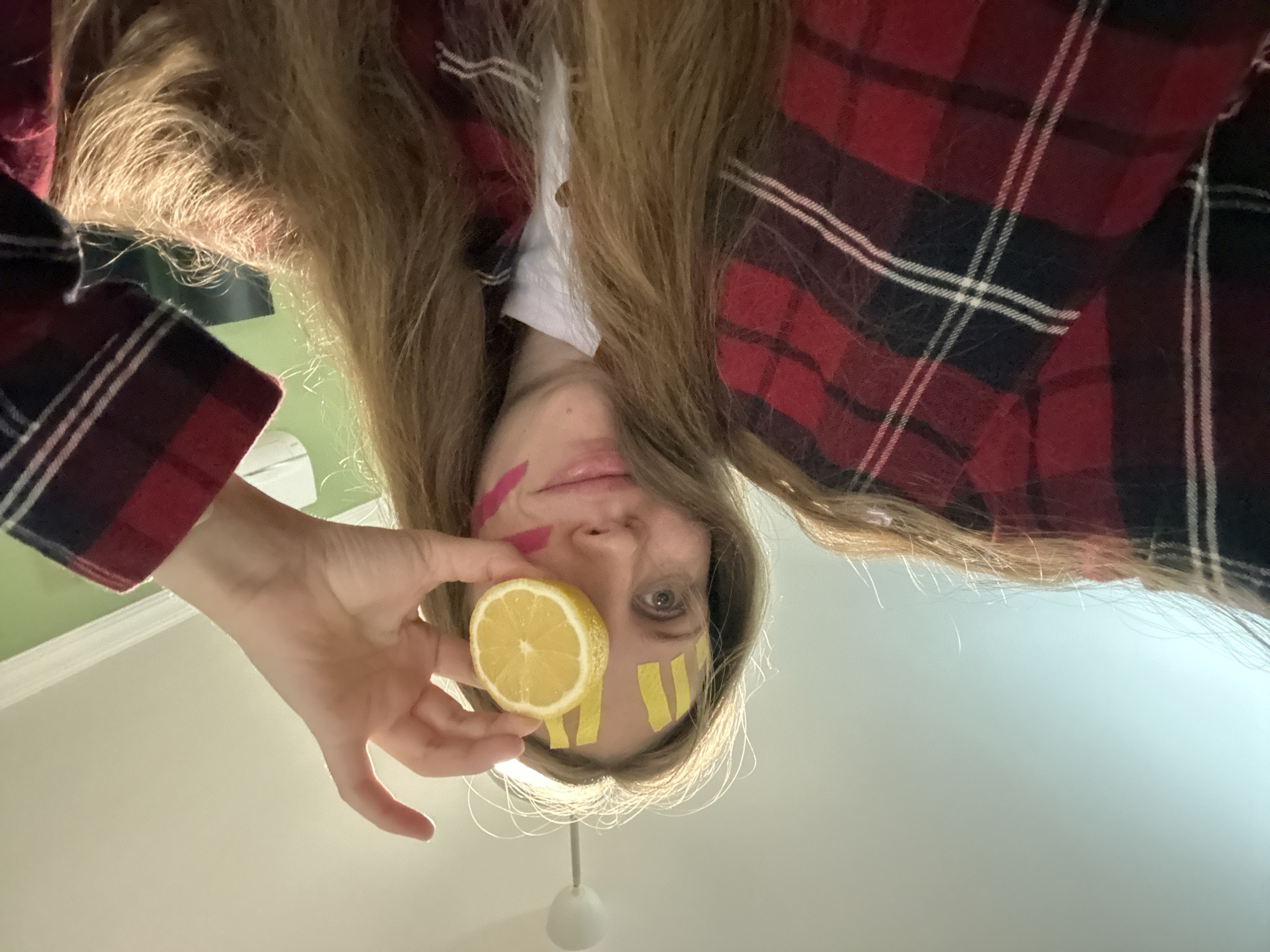 The image depicts a person holding a lemon slice in front of their face. The person has long, light brown hair and is wearing a red and black plaid shirt. There are yellow strips of tape on the person's face around the right eye and a pink mark on the left cheek. The background is a light gray ceiling with a light fixture.
The image does not provide any information about the location being Fethiye, Turkey.