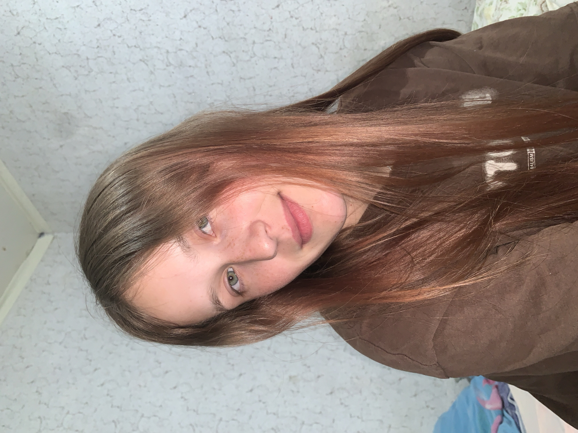A close-up portrait shows a young person, likely female, with long, reddish-brown hair, green eyes, and freckles on their nose and cheeks. They are wearing light pink lipstick and look directly at the camera with a subtle, soft smile and a slight head tilt.
The subject is dressed in a dark brown sweatshirt or jacket. On the garment, a white rectangular label-like element with black text is visible, partially obscured, but the word "MALISM" and parts of other letters suggesting "REALISM" can be discerned.
The background features a light-colored, possibly off-white or light gray, wall with a distinct, repetitive textured or patterned surface. In the upper right corner of the main image, a small portion of a bed or pillow with a floral-patterned sheet is visible. The indoor lighting appears soft and even. The activity captured is a casual portrait pose. The location context for this media file is Фоки, Russia.