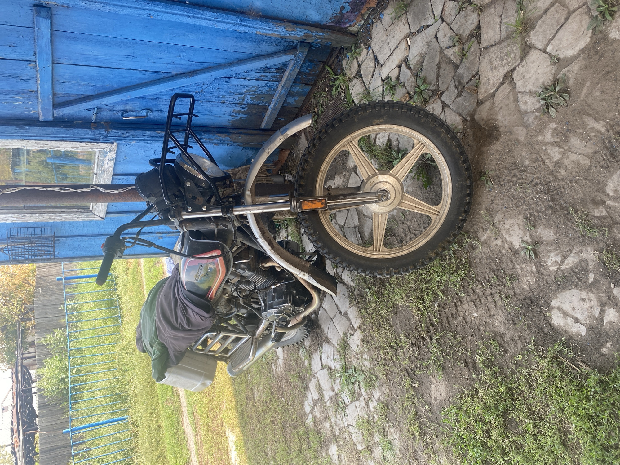 A motorcycle is parked in front of a weathered blue wooden door. The motorcycle is black and chrome, with a front rack and a white container strapped to the back, covered by a dark blanket and a green tarp. The motorcycle's front wheel, with a distinctive star-patterned rim, is prominent in the foreground. The ground is a mixture of dirt and irregular stone paving, with patches of grass. Behind the motorcycle, a light blue metal fence encloses a grassy area. In the background, a rustic building with a corrugated roof and a wooden fence can be seen. The scene appears to be in a rural setting, possibly a yard or driveway, within the Луганск ауылдық округі, Kazakhstan.