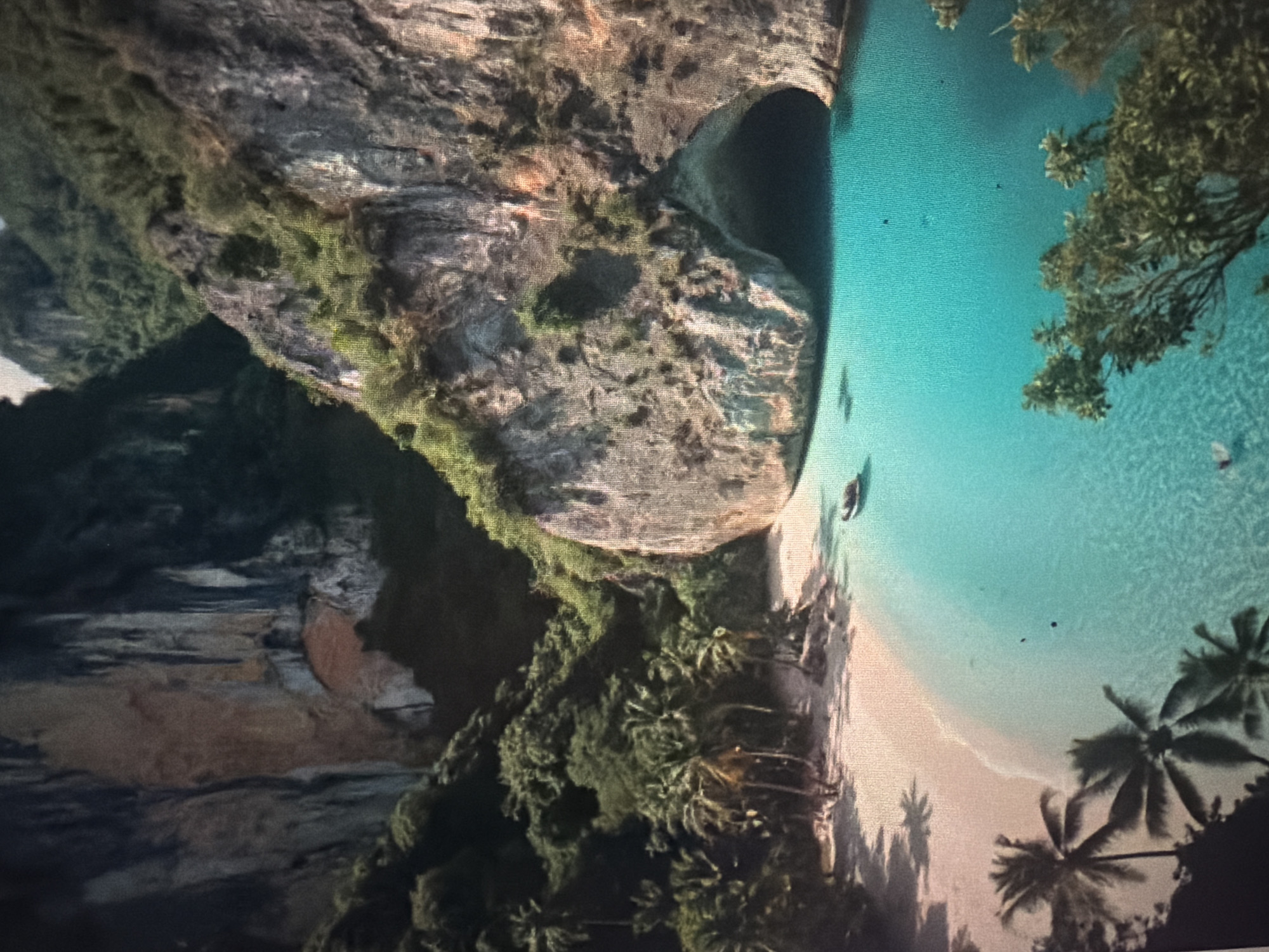 The image displays a scenic view of a tropical cove with turquoise waters, a sandy beach, and lush vegetation clinging to steep cliffs. The cove is characterized by a prominent cave opening in the rock face, suggesting a natural geological formation. Palm trees line the shore, indicating a warm, humid climate. A small boat is visible in the water near the beach, and what appear to be two individuals are swimming in the shallow sea. The overall impression is one of a secluded, idyllic paradise. The image does not provide any direct visual information that would allow for definitive identification of the location as Baan Mae Nam, Thailand.