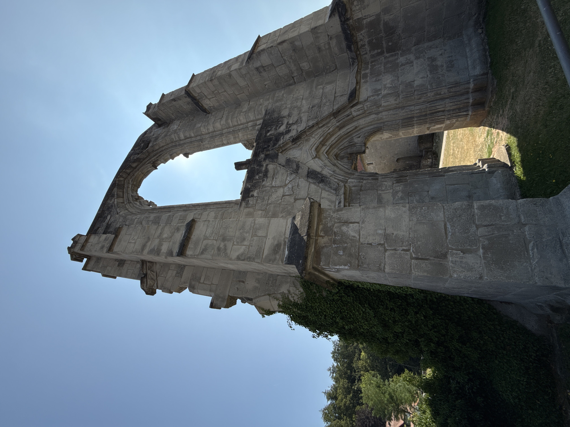 The image shows a low-angle view of the ruins of the Walkenried Abbey in Walkenried, Germany. The structure is made of large, light-grey stone blocks and features Gothic-style architecture with pointed arches and ornate detailing, though much of it is weathered and broken. Sunlight streams through a large archway in the center of the frame, creating a lens flare. Ivy clings to the left side of the stone structure. In the background, lush green trees and a hint of a building with a red roof can be seen. The ground in front of the ruins is covered in dry grass and some patches of green grass, with a shadow cast by the architectural remains. The sky is a clear, bright blue.