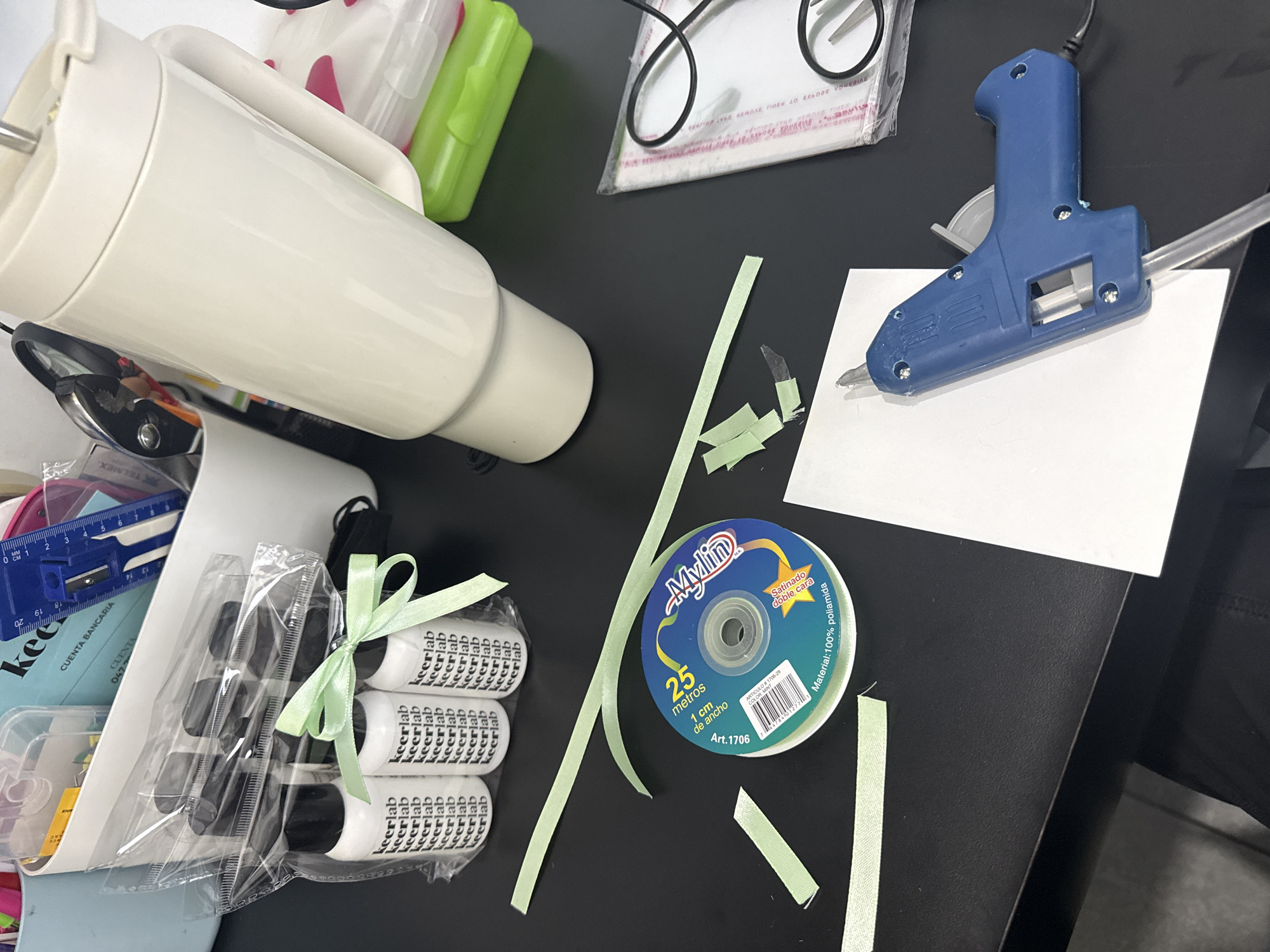 The image presents a cluttered black surface, likely a desk or table, scattered with various crafting and office supplies. A white electric device, possibly a food processor or blender base, takes up space on the left side. Next to it is a clear plastic package containing three white bottles labeled "keerlab" and a small blue measuring tool. A spool of light green ribbon, labeled "Mylin," is positioned near several cut pieces of the same ribbon. A blue glue gun rests on a white square of paper on the right side of the surface. Other items visible include a clear plastic bag with a black cord, a green container, and various pieces of paper and stationery. The context of San Bernardino Tlaxcalancingo, Mexico is not evident from the image itself.