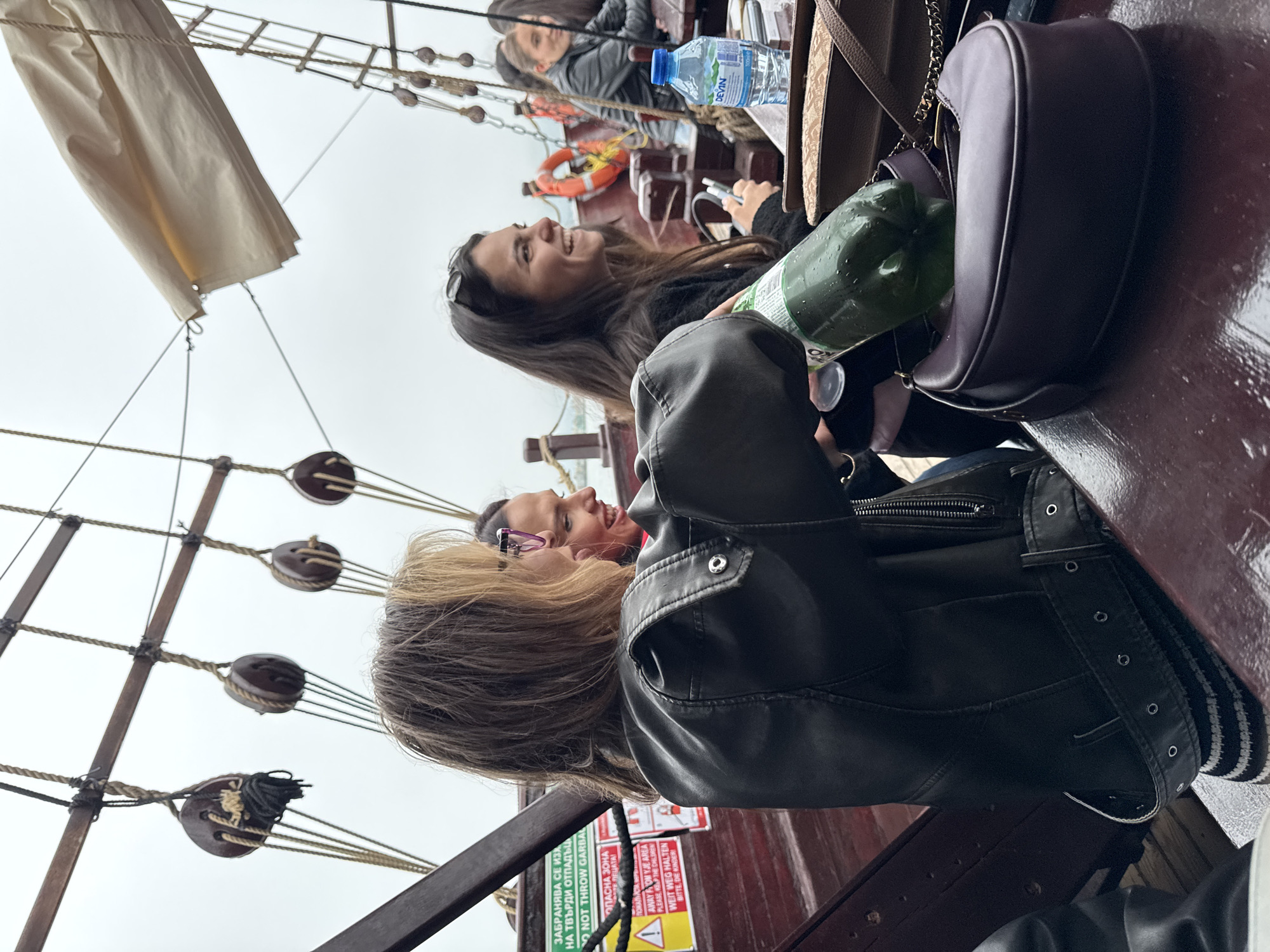 Two women are seated and smiling on a boat, potentially a replica pirate ship, with traditional rigging visible. A life preserver and water bottle are on the table behind them. A warning sign is visible on the side of the boat. Based on the context clues, the location may be Varna, Bulgaria.