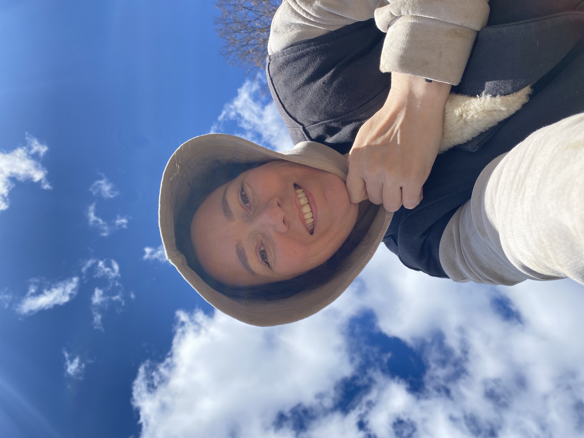 A woman with a light complexion and a warm smile faces the camera, her face framed by a light brown bucket hat. She is wearing a black jacket with beige sleeves and a fuzzy white interior. Her hand is visible, gently touching her cheek. The background is a bright blue sky dotted with fluffy white clouds. In the top center of the frame, a bare tree branch can be seen. The photograph is taken from a low angle, looking up at the subject. The location appears to be outdoors, under clear skies. The location, Ольховка, Russia, is not discernible from the image itself.