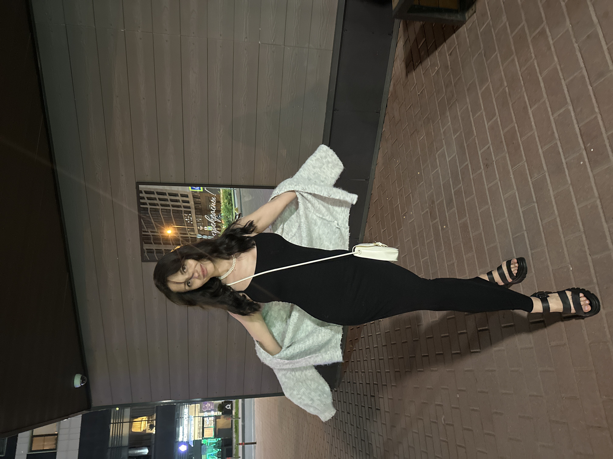 The image depicts an adult female standing outdoors at night in an urban environment in the city of Unknown, Unknown. She is positioned slightly to the left of the frame's center, facing the camera with a faint smile. She has long, dark brown hair, some strands appearing windblown. She is wearing a black, form-fitting, sleeveless maxi-dress with spaghetti straps, a pearl necklace, and black chunky platform sandals with multiple straps and a buckle. A light grey or off-white knit cardigan is draped loosely over her shoulders, with the sleeves partially covering her arms. A small white cross-body bag with a thin strap hangs on her left hip (viewer's right).
The woman stands on ground paved with reddish-brown rectangular brick pavers. Behind her, to the right, is a building facade featuring horizontal grey-brown siding panels above a darker grey-black base. A framed, reflective surface on this wall displays a blurred urban scene and partially legible Cyrillic text, including "прекрасны!" (beautiful!). Above this, a white dome-shaped security camera is mounted on the underside of an overhang. To the left of the woman, and in the distant background, is a modern multi-story building with numerous illuminated windows. Further down the street, various illuminated storefronts emit green and purple light, and streetlights are visible. A dark, possibly metal, bench or planter is partially visible in the far right foreground. The scene is illuminated by artificial night lighting, casting shadows on the ground.