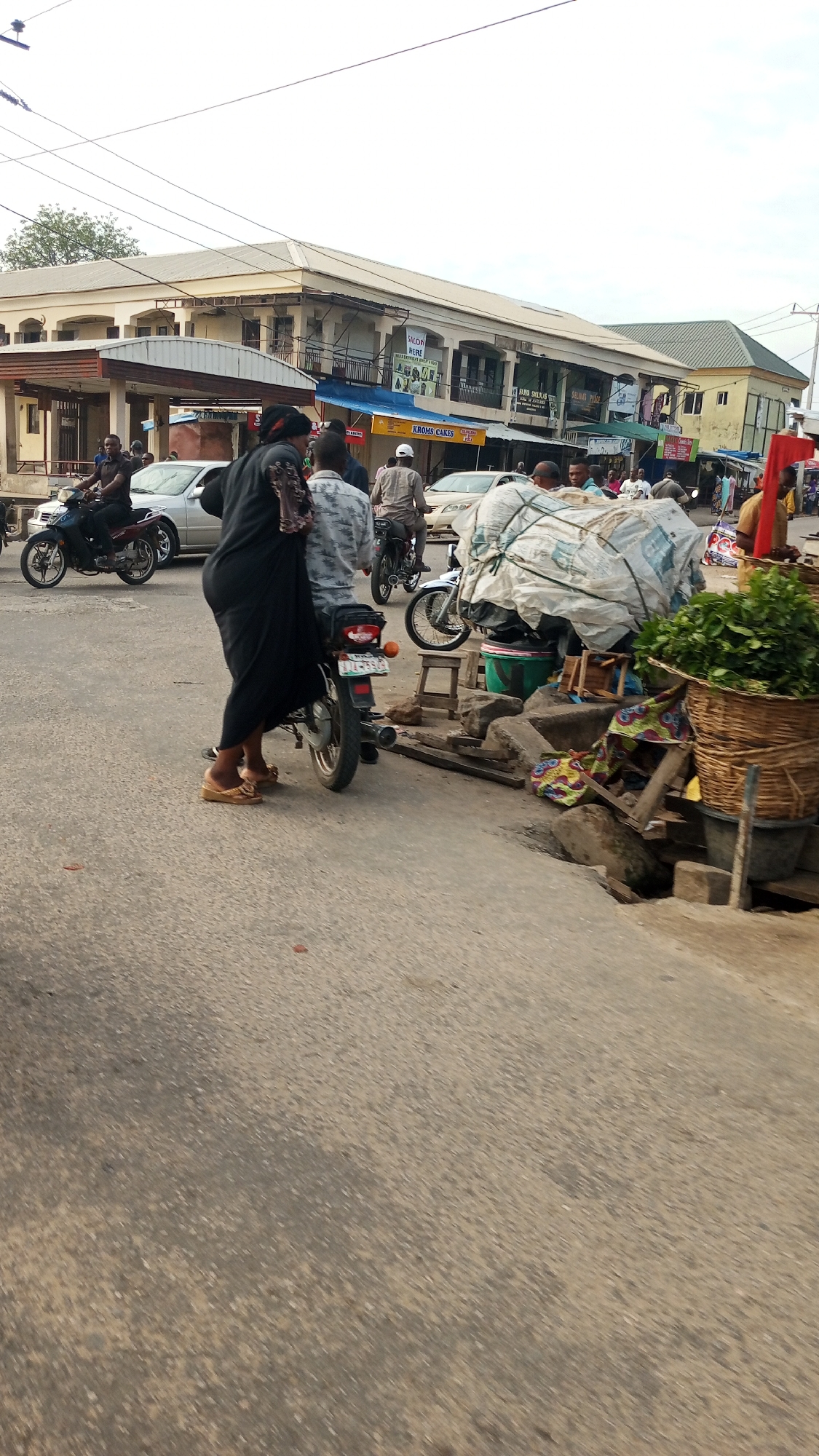 The image depicts a busy street scene in Lokoja, Nigeria, during daylight hours under a bright sky. The setting appears to be a commercial or market area, characterized by multi-story buildings housing various shops and bustling activity on the street.
In the immediate foreground, a woman dressed in a black long dress and wearing brown sandals is standing beside a black motorcycle, which has a visible license plate: LJK-853-QT. Another individual is partially visible on the motorcycle behind her. To the right, a makeshift market stall is set up along the roadside, featuring a large woven basket filled with green leafy vegetables. Adjacent to it are various goods, some covered by a white tarpaulin or sheet, stacked on concrete blocks and a small wooden stool above a visible drainage ditch.
The midground and background are filled with people and vehicles. Several motorcycles, commonly known as "okadas," are prominent, with riders and passengers navigating the street. A silver car is also visible in the traffic. People are walking on the pavement and alongside the road, contributing to the lively atmosphere.
The buildings lining the street are mostly two to three stories high, with shops occupying the ground floor. Several storefront signs are discernible: "SALON BIEN" with "BEST HAIR SALON & GIFT SHOP" underneath, "KROMS CAKES," "NUR STUDIO," and "SELMA'S PLACE." Overhead power lines crisscross the frame against the bright sky. The road is paved with asphalt, and a concrete curb separates it from the pedestrian area and market setup. The scene captures the daily vibrant street life and commerce typical of a Nigerian town.
