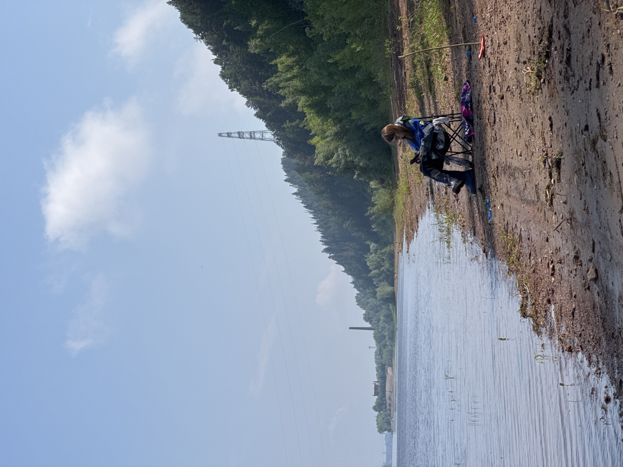 A person sits in a folding chair on a sandy shore next to a body of water. The person is wearing a blue jacket, dark pants, and boots, and is looking down at a mobile device. A colorful blanket is draped over the back of the chair. To the right of the chair, a long stick is propped up in the sand with a red object at its base. The sandy shore slopes up to a dense forest of green trees, with a tall, lattice-style power pylon visible in the distance on the hillside. The sky is a pale blue with a few wispy clouds. In the far distance, on the left side of the image, there are buildings and a tall smokestack. The water is calm with ripples.