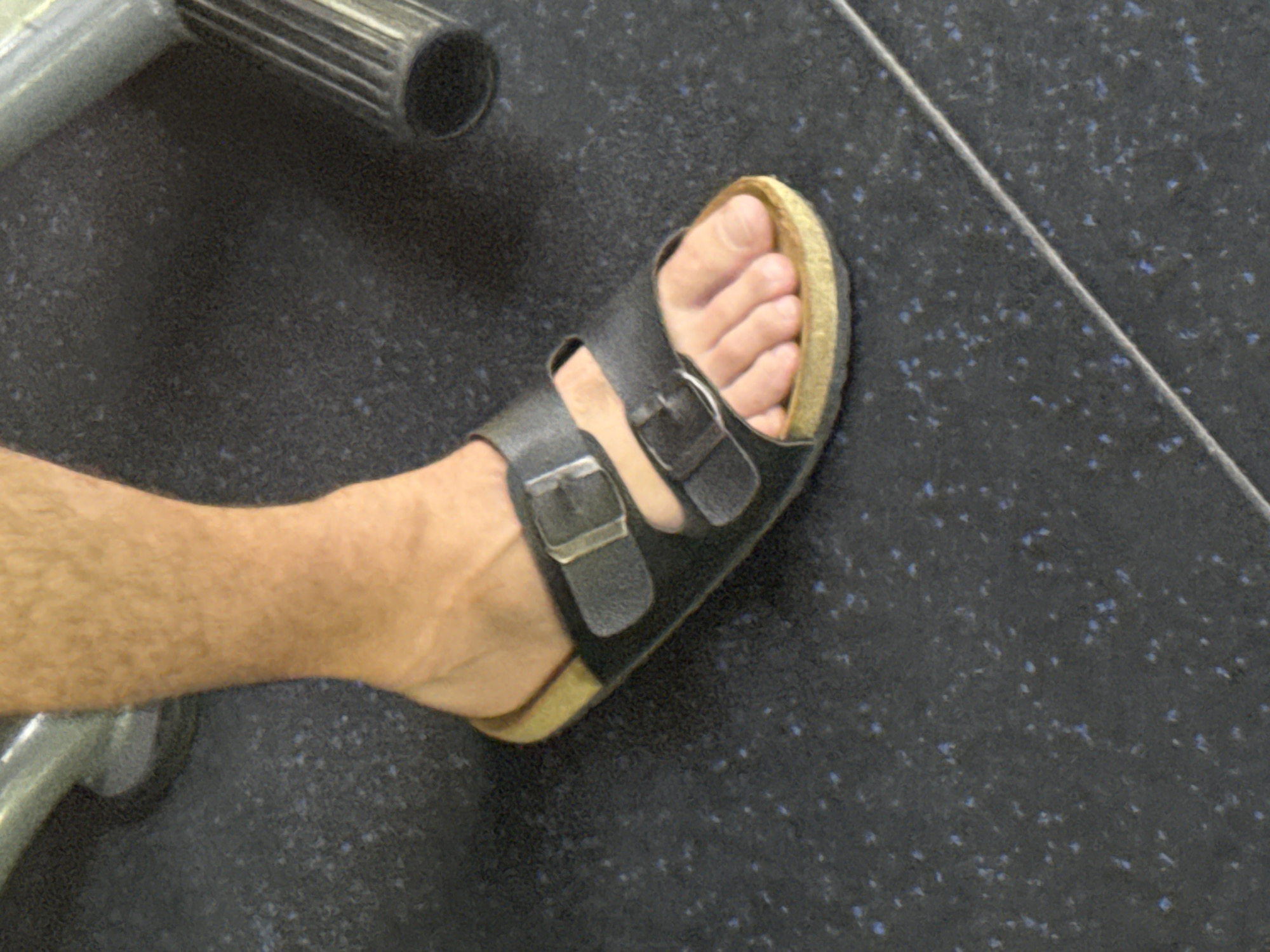 The image shows a person's foot wearing a black sandal with two straps and buckles. The sandal has a cork footbed. The foot is on a dark gray floor with small blue speckles. In the background, a portion of a metallic object, possibly part of a vehicle or gym equipment, is visible. There is no information present to suggest this photo was taken in Canggu, Indonesia.