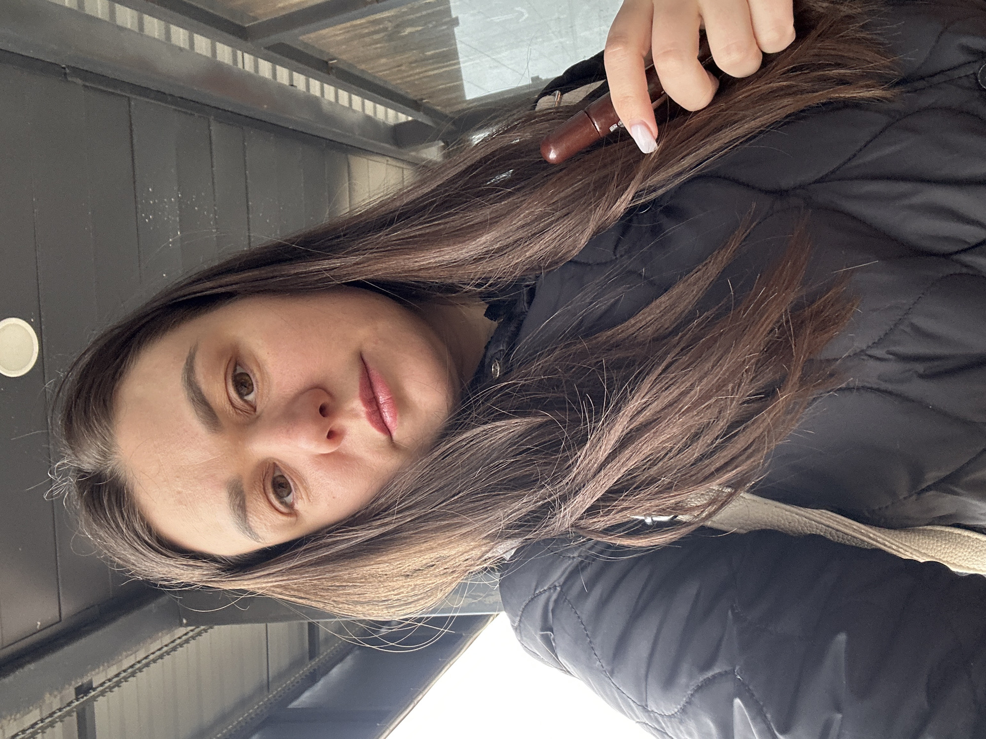 A woman with long brown hair is holding a brown lip pencil in her right hand. She's wearing a black puffer jacket with a cream-colored collar. The location appears to be in Chaikovsky, Russia, based on the background elements.