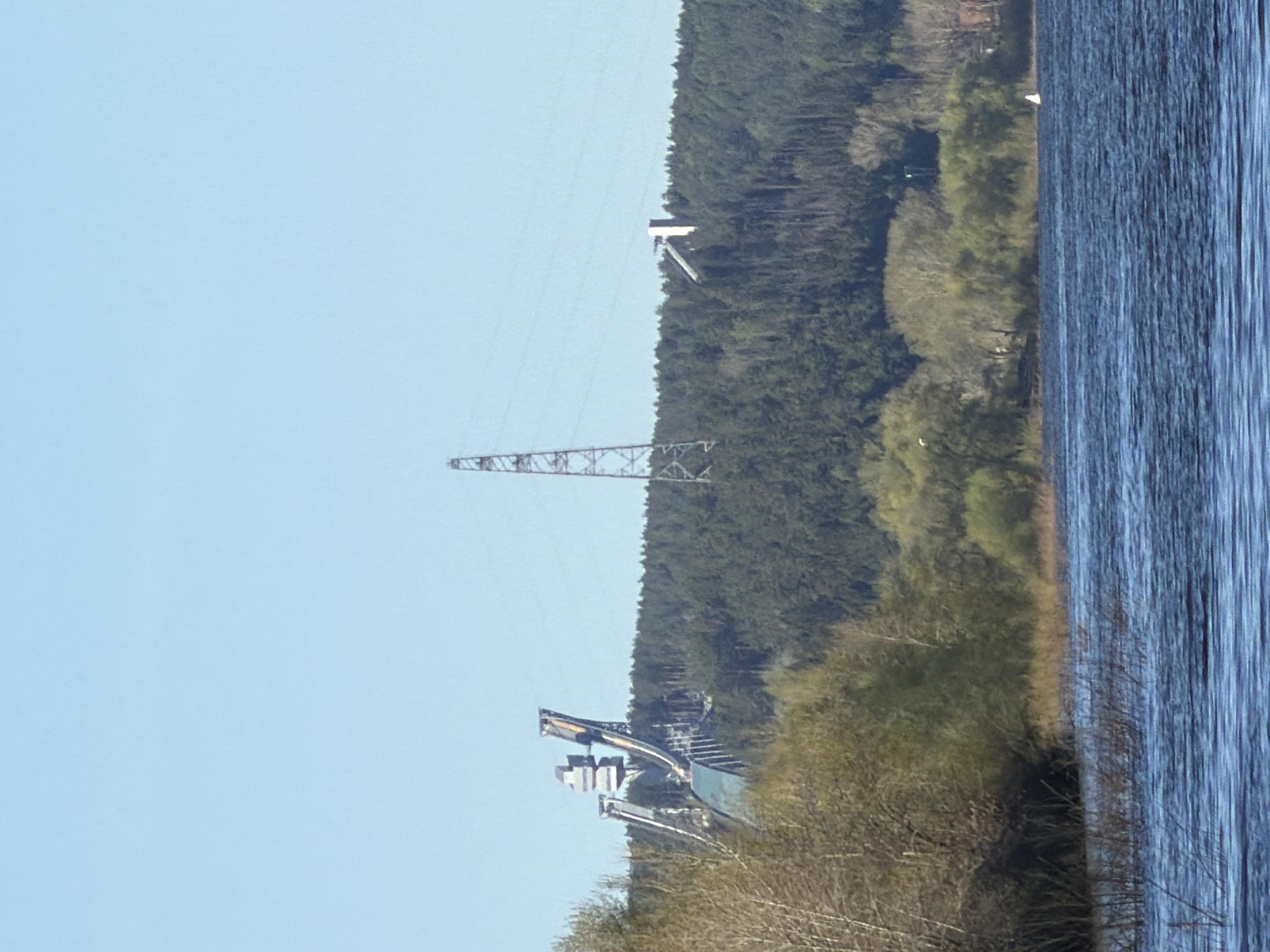 The image shows a view of the Votkinsk Hydroelectric Station in Chaikovsky, Russia. A body of water, possibly the Votkinsk Reservoir, is on the right. Adjacent to the water is a tree-covered area, likely part of the surrounding landscape. Power lines run parallel to the landscape. The hydroelectric station, with its infrastructure, is visible in the middle of the image against the backdrop of trees. The sky is clear and blue.