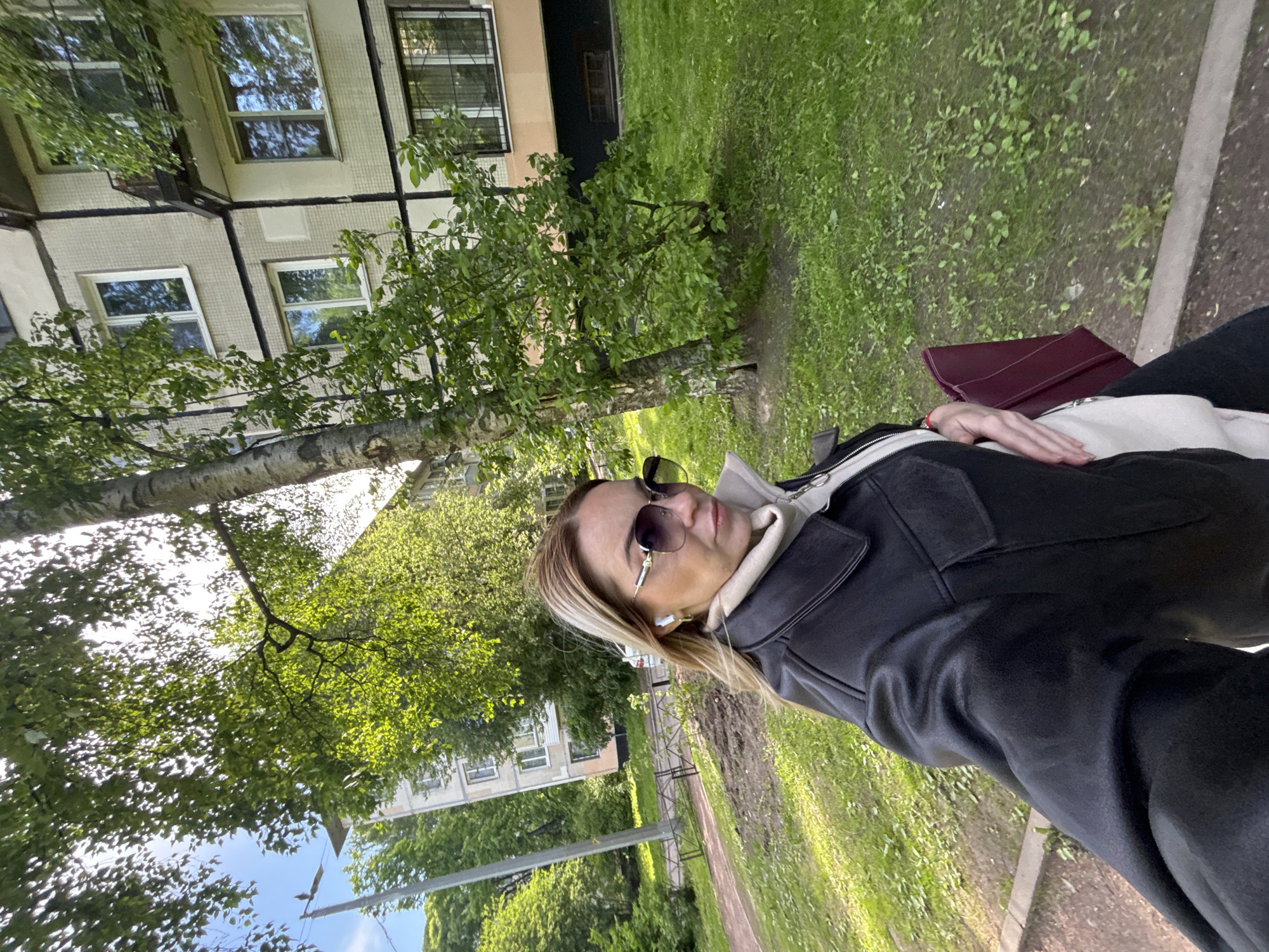 The photograph captures a woman seated on what appears to be a bench in a grassy area. Buildings typical of Soviet-era construction are visible in the background, suggesting a location in a residential district possibly within Saint Petersburg, Russia. The woman is wearing sunglasses and a dark jacket. A burgundy-colored handbag rests on her lap. The scene is outdoors with the presence of trees and greenery.