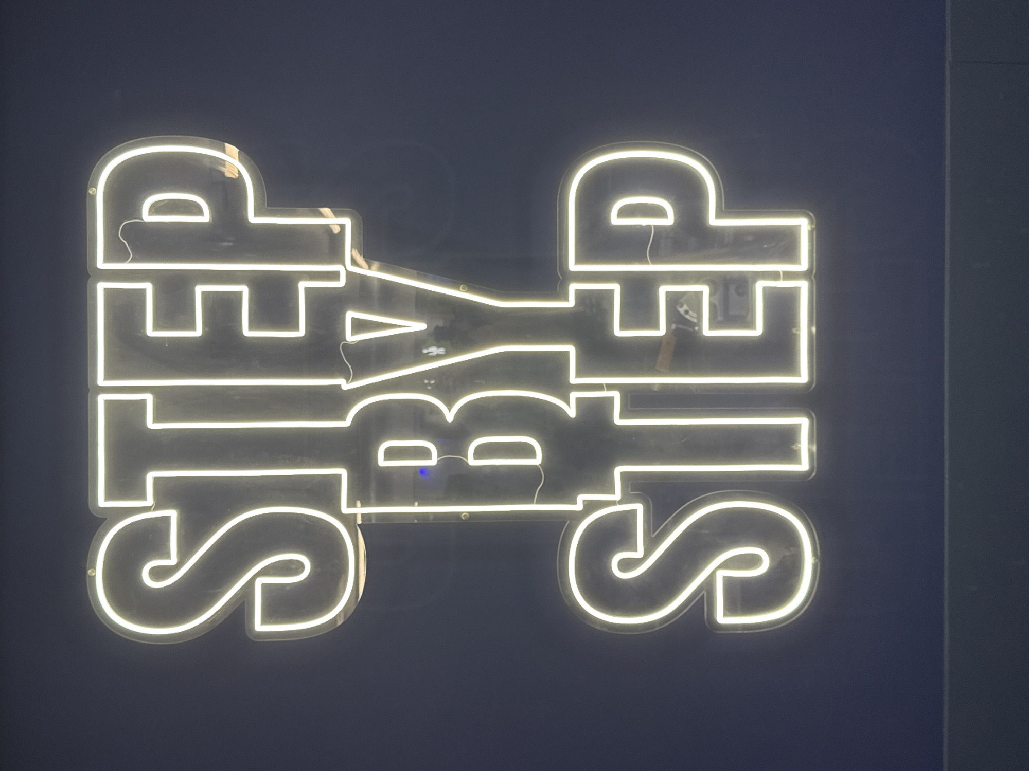 A neon sign, glowing with bright, cool white light, is prominently displayed against a dark, plain wall, likely deep blue or charcoal gray. The sign consists of two words arranged vertically. The bottom word, "STRIPE," is presented right-side up in a bold, stylized block font with rounded edges. Directly above it, the word "STREETS" is rendered in the same font but is inverted (rotated 180 degrees), creating a visually striking and symmetrical effect.
The illumination from the neon tubes strongly contrasts with the dark background, suggesting the photograph was taken in low light conditions, possibly indoors or at night in an urban setting such as London, United Kingdom. Fine details of the sign's construction are faintly visible, including the clear acrylic or glass backing supporting the neon tubes, thin connecting wires, and small mounting screws. A tiny blue indicator light can be seen near the center of the sign, hinting at its electrical components. No people or other objects are present, ensuring the neon artwork remains the sole focus of the image.