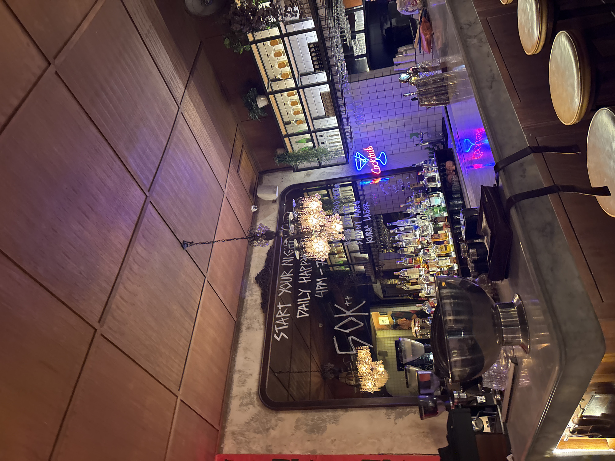 The image depicts the interior of a dimly lit bar, with warm wooden paneling on the left and a marble bar counter on the right. The setting appears to be a trendy establishment, possibly a cocktail bar, as suggested by a neon "Cocktails" sign and a wall adorned with bottles of liquor.
The focal point in the foreground is a large, ornate mirror reflecting the bar's ambiance. Written in chalk on the mirror are the words "START YOUR NIGHT", "DAILY HAPP", and "4PM-7PM". Below this, "50K++" is also visible in chalk, likely indicating a price or special offer. Several chandeliers with intricate designs are visible both in reality and in the reflection, adding a touch of elegance.
Behind the bar, shelves are packed with various bottles of spirits and liqueurs, illuminated by a blue neon sign. The overall atmosphere is one of sophisticated nightlife, with subtle lighting creating a mood conducive to relaxation and socializing. There are no people clearly visible in the frame, but the setting suggests it is open for business, potentially during happy hour. The location context of North Kuta, Indonesia, places this bar within a popular tourist and entertainment area.