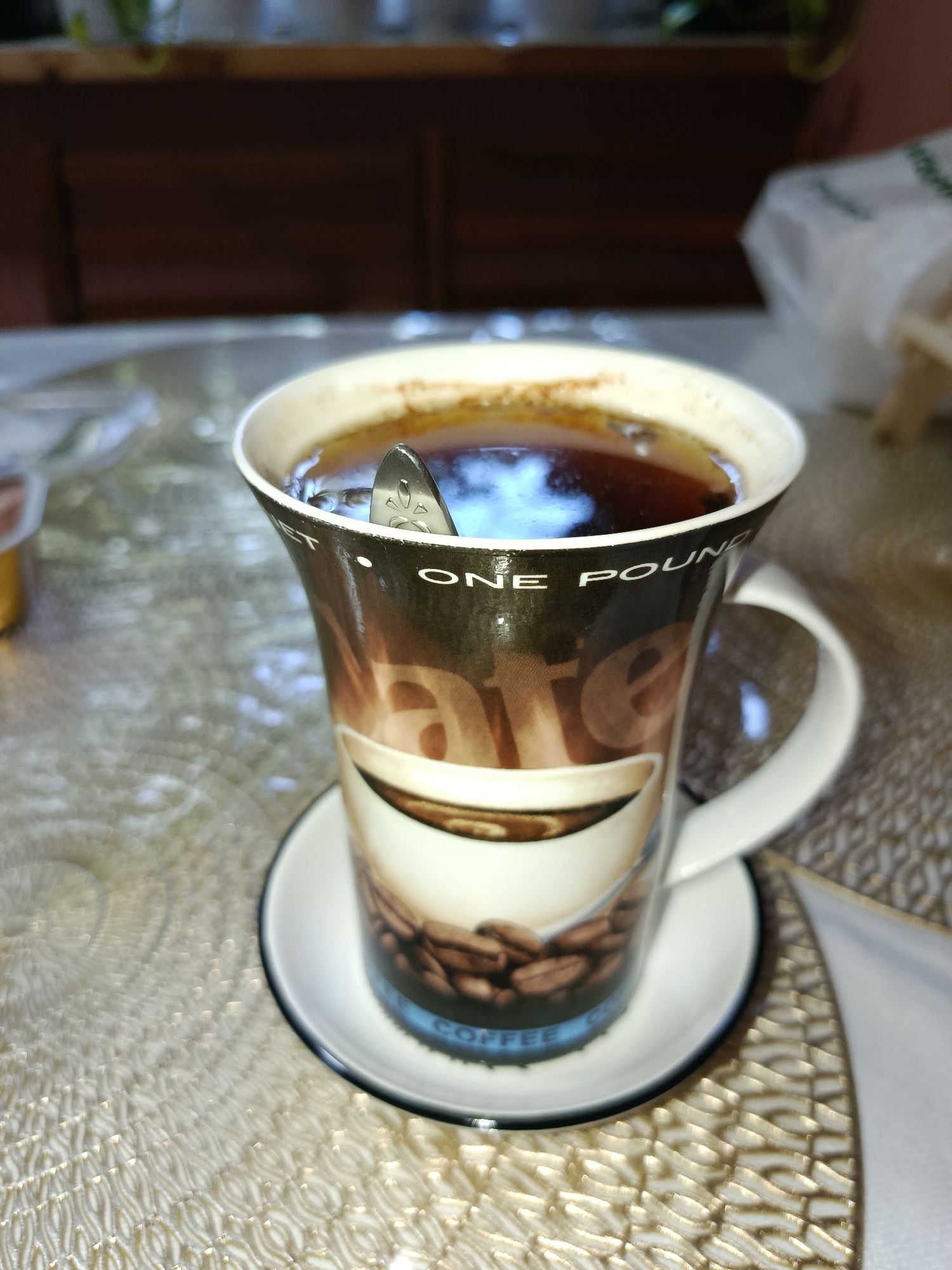 A close-up, eye-level shot shows a coffee mug on a saucer, placed on a textured, light-colored tablecloth. The mug is filled with dark liquid, likely coffee, and a silver spoon rests partially submerged within it. The mug features a graphic design with the words "ONE POUND" and an image of coffee beans. The background is out of focus, revealing hints of wooden furniture and possibly other objects on a shelf. The lighting suggests an indoor setting, possibly a home or cafe. The scene is static with no discernable activity. The location of this image is not specified to be in Tashkent, Uzbekistan.