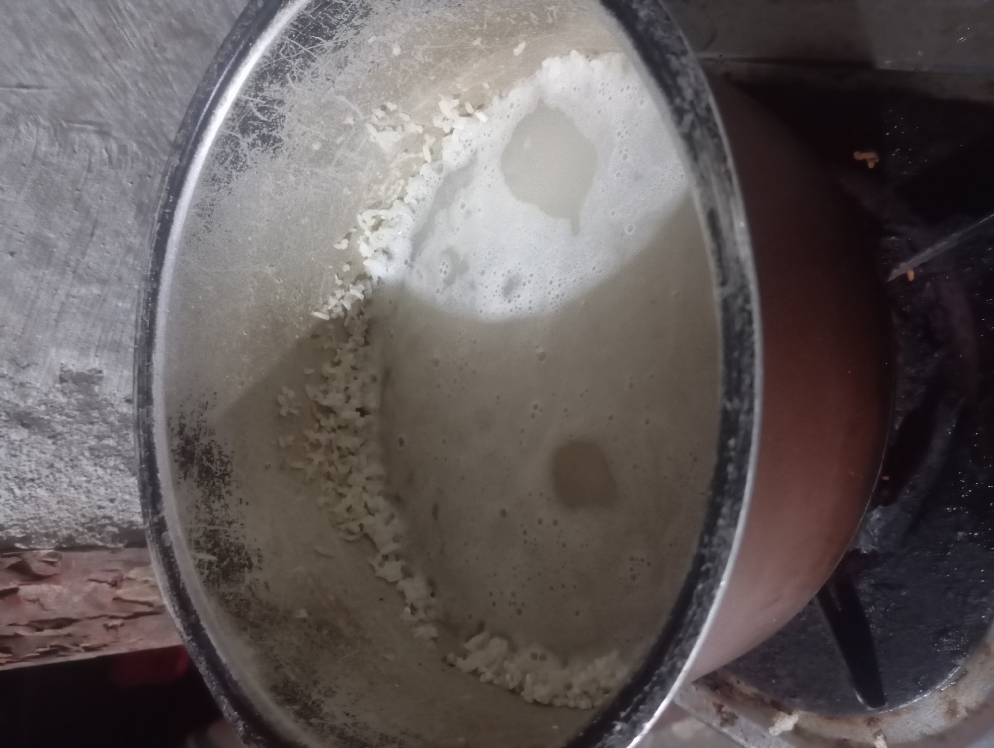 A close-up shot shows a pot of rice cooking on a stovetop. The pot is made of metal and has a brown exterior. The rice is in the process of cooking, with some grains already soft and submerged in the starchy water, while others cling to the sides of the pot. The water is foamy and milky, indicating that the rice is releasing starch. The pot is placed on a black metal stove burner, with some food debris visible on the burner and around the base of the pot. The background is out of focus, but appears to be a rough, textured wall. The scene is lit by an overhead light source, casting shadows and highlighting the texture of the food and the environment. The image does not contain any information about a specific location in the city of Dente Teladas, Indonesia.