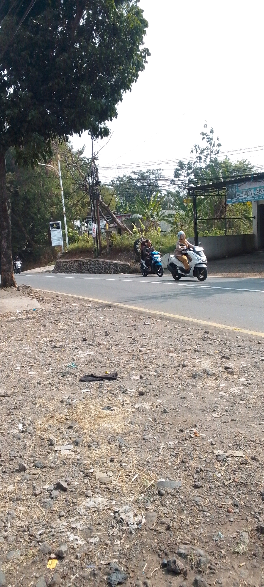 A man and a woman ride on a scooter along a paved road in Cilaku, Indonesia. The woman in front is wearing a light brown hijab and a yellow shirt, while the man behind her is wearing a dark blue shirt. They are traveling on a road with a yellow line marking the shoulder. In the background, there are trees and buildings, including a sign that reads "JUAL BA...". The ground beside the road is covered in gravel and debris. There is another scooter with one rider in the distance on the left side of the road.