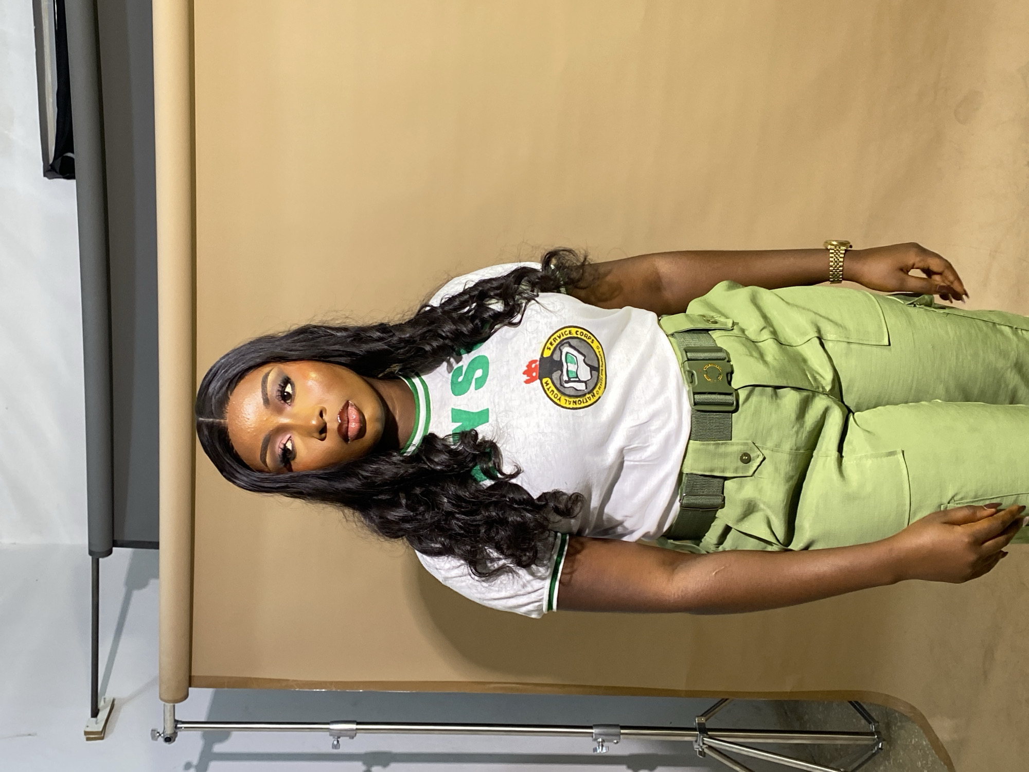 A young Black woman stands in a studio setting against a tan backdrop. She has long, dark, wavy hair and is wearing a white t-shirt with green trim and a graphic on the chest, and light green cargo pants with a matching belt. Her makeup is visible, with dark eyeliner and eyeshadow. She is looking to her right, away from the camera. In the background, there are studio lighting stands and rolled backdrops. The image does not provide any specific details about a location in Gura Top, Nigeria.
