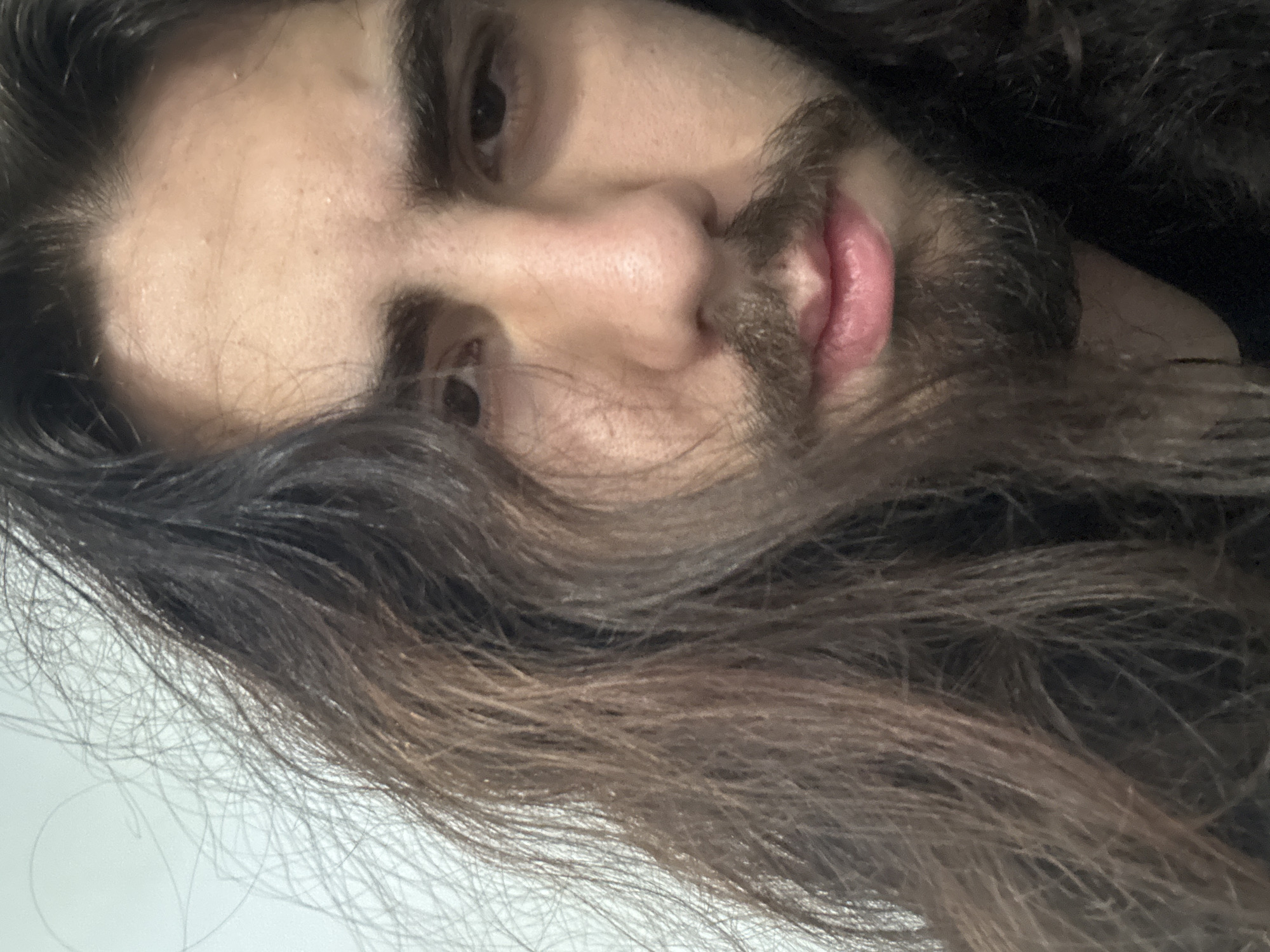 A close-up, shoulder-up, eye-level, medium shot shows a young male with long, dark, wavy hair and a beard. He is looking forward with his mouth slightly ajar. The background is a plain, light-colored wall.