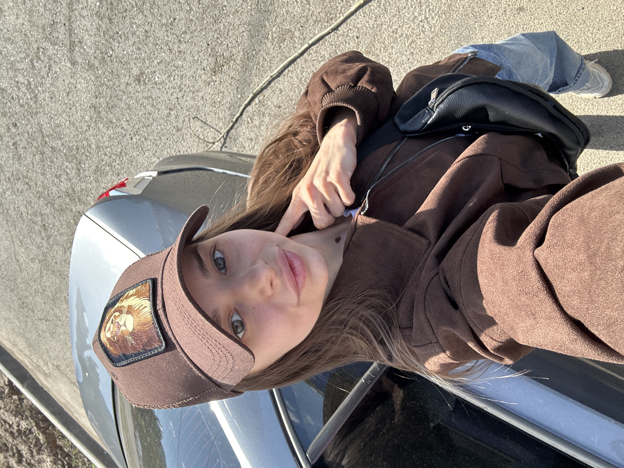 A young woman is sitting on a silver car, possibly in Izhevsk, Russia. She is wearing a brown cap with a lion patch, a brown jacket, and jeans, and she has a black bag across her shoulder. The car is parked on pavement. The photo is taken from a high angle.