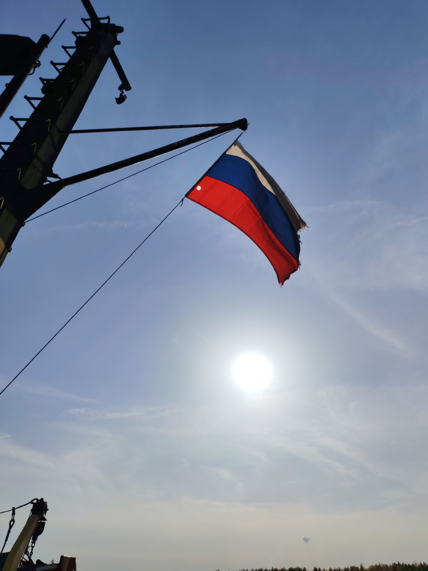 A tattered Russian flag hangs from a metal arm extending from a dark, possibly metallic, structure, potentially part of a ship or industrial equipment. The flag's colors (white, blue, red) are clearly visible. The background is a bright, hazy sky with the sun shining directly behind the flag. The scene appears to be outdoors. A thin cable runs diagonally from the structure to the area near the sun. The bottom portion of the image suggests the edge of some equipment and trees in the distance, perhaps denoting a coastal area. No people are visible in the frame. The location is specified as Shelomovo, Russia.