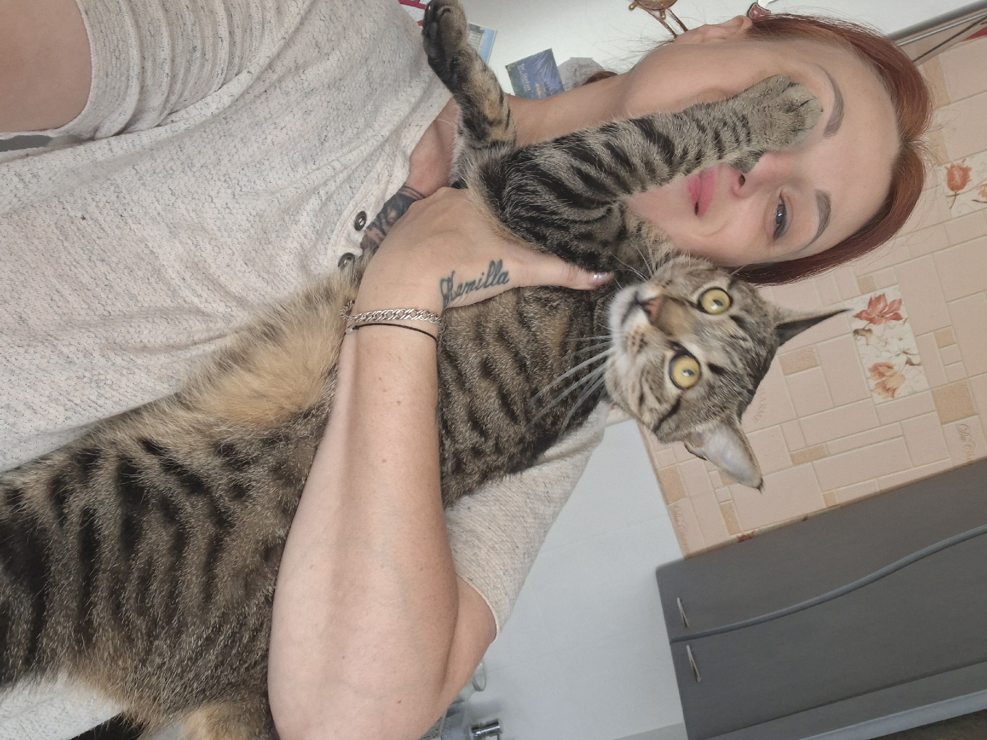 The image shows a woman holding a tabby cat. The woman has red hair and is wearing a light-colored, textured shirt. Her left arm is raised, with the cat's paw resting on her face. The cat, with yellow eyes and striped fur, is looking to the right. The woman has a tattoo on her left forearm and is wearing a bracelet. The background appears to be an indoor setting with a tiled wall and a grey cabinet. There are no discernible landmarks or elements in the image that would suggest a specific location within the city of Myadzyel, Belarus. The scene is a casual, close-up shot.