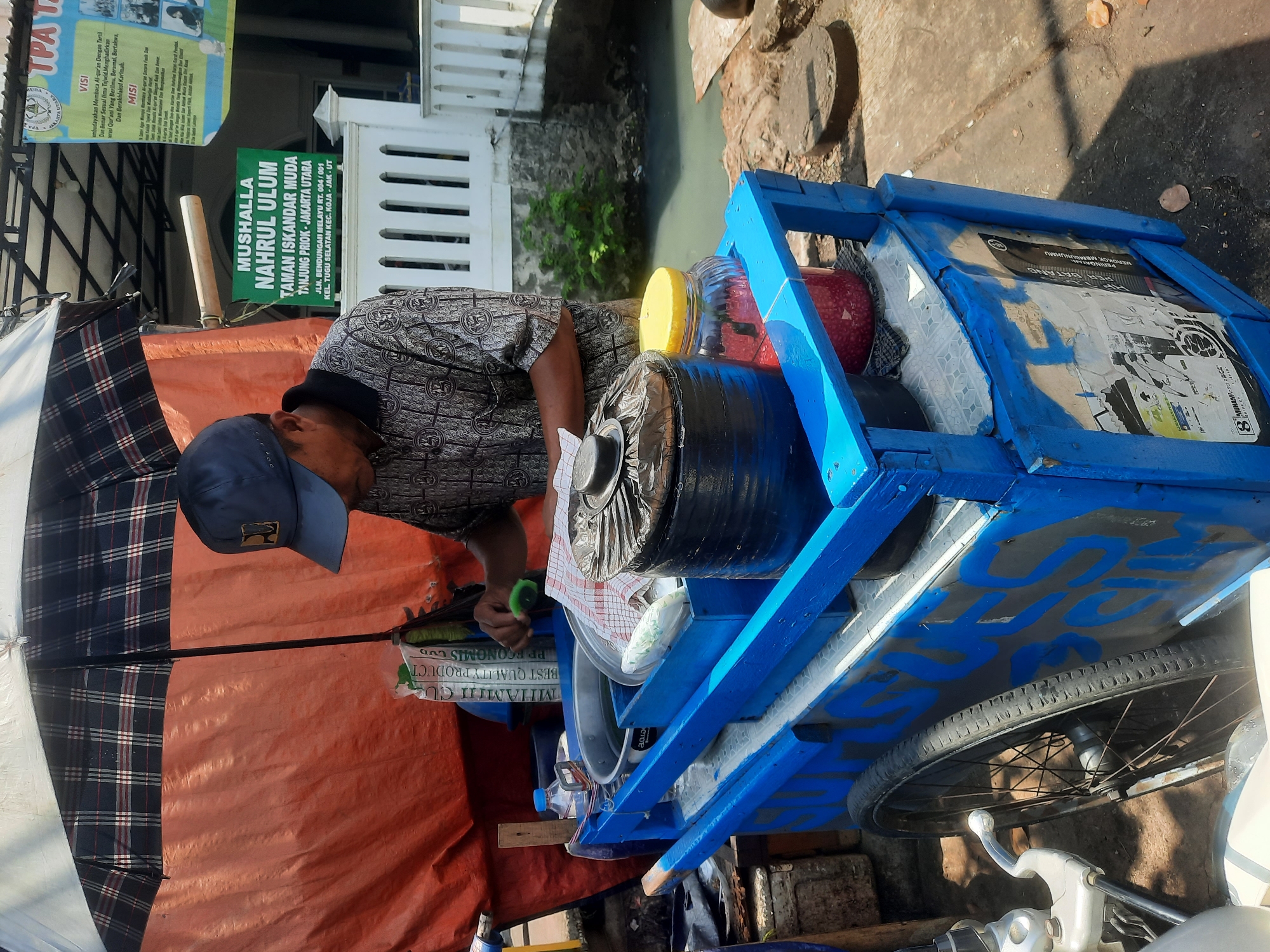 A man wearing a blue baseball cap and a patterned shirt operates a blue food cart under a plaid umbrella. He is standing on a sunny paved area, possibly a sidewalk or street, next to a canal. The food cart is filled with various containers, including a large metal pot and a glass jar filled with red beads. A green sign behind the man reads "MUSHOLLA NAHRUL ULUM" and "TANJUNG PRIOK - JAKARTA UTARA," indicating the location as Tanjung Priok in North Jakarta, Indonesia. The man is in the process of preparing food, holding a small green object in his right hand. The background shows a white railing and some vegetation. The overall scene depicts a street vendor in a bustling urban environment.