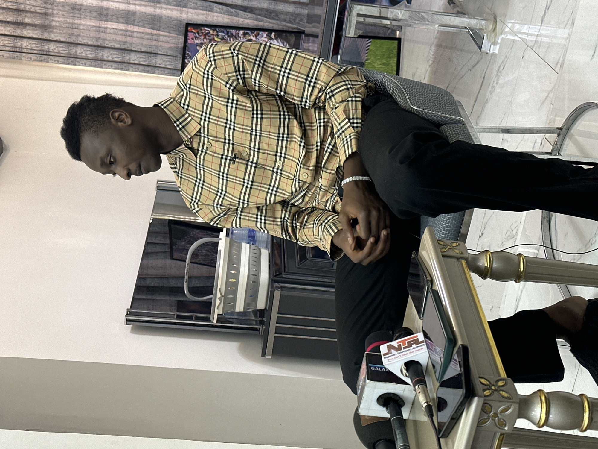 This image captures a young Black man sitting in what appears to be an indoor setting, possibly a press conference or interview. He is wearing a long-sleeved, beige plaid shirt with black and red accents, black pants, and a silver diamond-studded bracelet on his left wrist. His head is bowed, and his hands are clasped in his lap, suggesting a moment of contemplation or shyness. The background includes a patterned curtain, a television screen displaying what looks like a sports event, and a modern chair with a grey textured seat.
In the foreground, several microphones are visible, indicating a media event. One microphone has the "NTA" logo, identifying it as belonging to the Nigerian Television Authority. Another microphone has the "GALAXY" logo. The presence of these microphones and the man's attire suggest he is the subject of an interview or press briefing. The floor appears to be tiled with a marble pattern. The lighting suggests it is daytime, and there are no indicators of weather conditions. The overall mood conveyed by the man's posture is one of introspection. The location context provided is Unguwar Sarki, Nigeria.