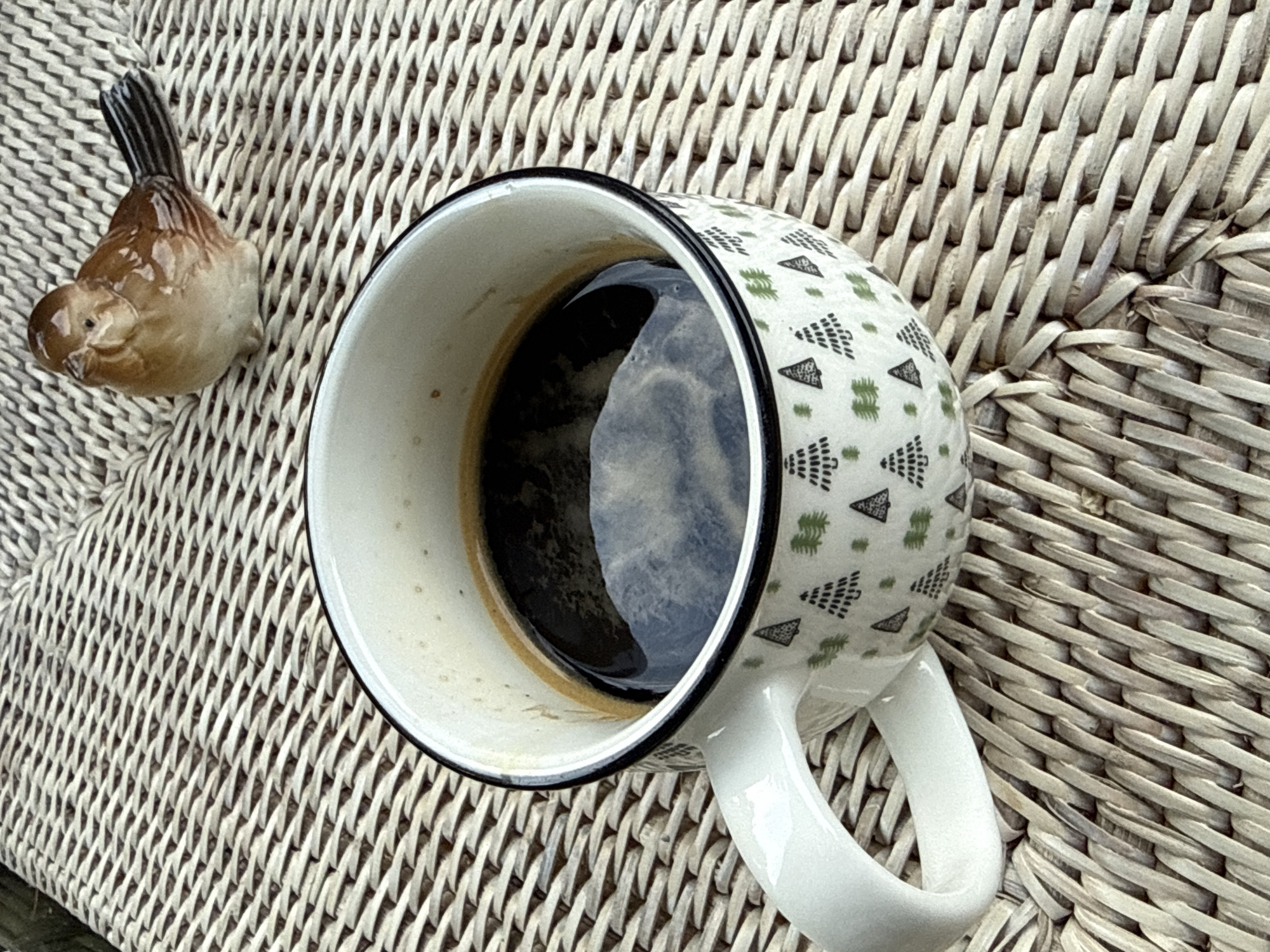 A close-up, eye-level shot captures a cup of coffee and a decorative ceramic bird on a woven surface. The mug, white with a black rim, is filled with dark coffee and sits slightly off-center to the left. It features a pattern of small green trees. To the upper right of the mug, a small, glossy ceramic bird with brown and white plumage is positioned. The background consists of a textured, light brown woven material, possibly a placemat or furniture. The scene is static and appears to be outdoors, given the natural lighting. No people or actions are depicted. The location in Bad Sachsa, Germany, is not discernible from the image itself.