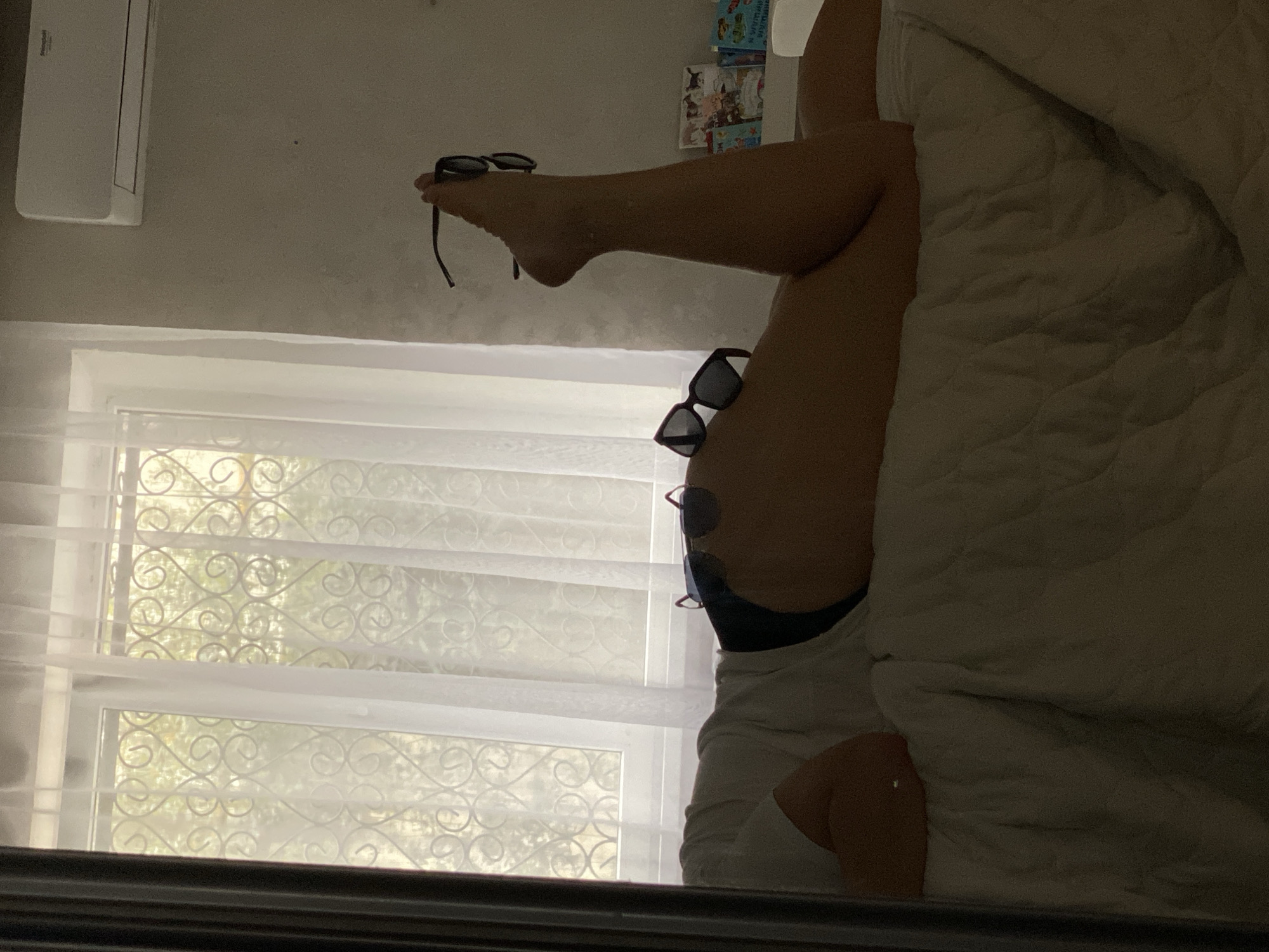 A person's legs are held up in the air, with sunglasses balanced on the foot. Another pair of sunglasses is balanced on the thigh. The person is wearing a white shirt and dark underwear. The background consists of a window with sheer curtains and a glimpse of greenery outside. A bed with a thick comforter is partially visible. The location appears to be indoors, likely a bedroom in a city like Sochi, Russia.