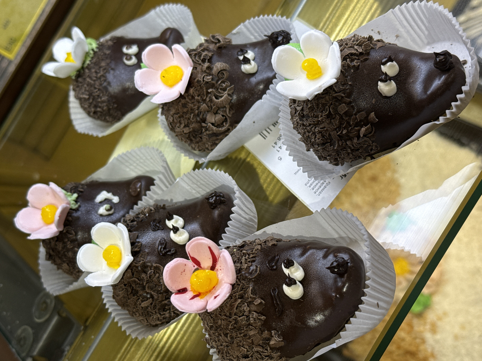 The image displays a close-up view of several whimsical chocolate desserts, likely cakes or pastries, presented in a display case. Each dessert is shaped like a small animal, resembling a hedgehog or similar creature, and is individually placed in a white fluted paper liner.
The desserts are coated in smooth, dark chocolate. One side of each, representing the "body" or "fur," is generously covered with dark chocolate shavings, creating a textured, spiky appearance. The smooth "face" portion of each dessert features two small white icing dots for eyes and a tiny dark chocolate dot for a nose or mouth, giving them a charming expression. Many of these chocolate creatures are adorned with delicate, edible sugar flowers on their heads or backs. These flowers are either light pink or white, both with a bright yellow center, adding a touch of color and decor.
The treats are arranged on a golden-yellow surface within what appears to be a glass display case, indicated by faint reflections and a glass divider. In the background, a piece of paper with text written in Cyrillic script is partially visible. Words such as "крахмал" (starch), "мука" (flour), and fragments of "яичное" (egg) and "сладкое" (sweet) can be discerned, along with numbers like "41" and "5." The scene suggests an indoor setting, likely a bakery or patisserie, showcasing these appealing, individually prepared items. The time of day and weather are not identifiable from the image.