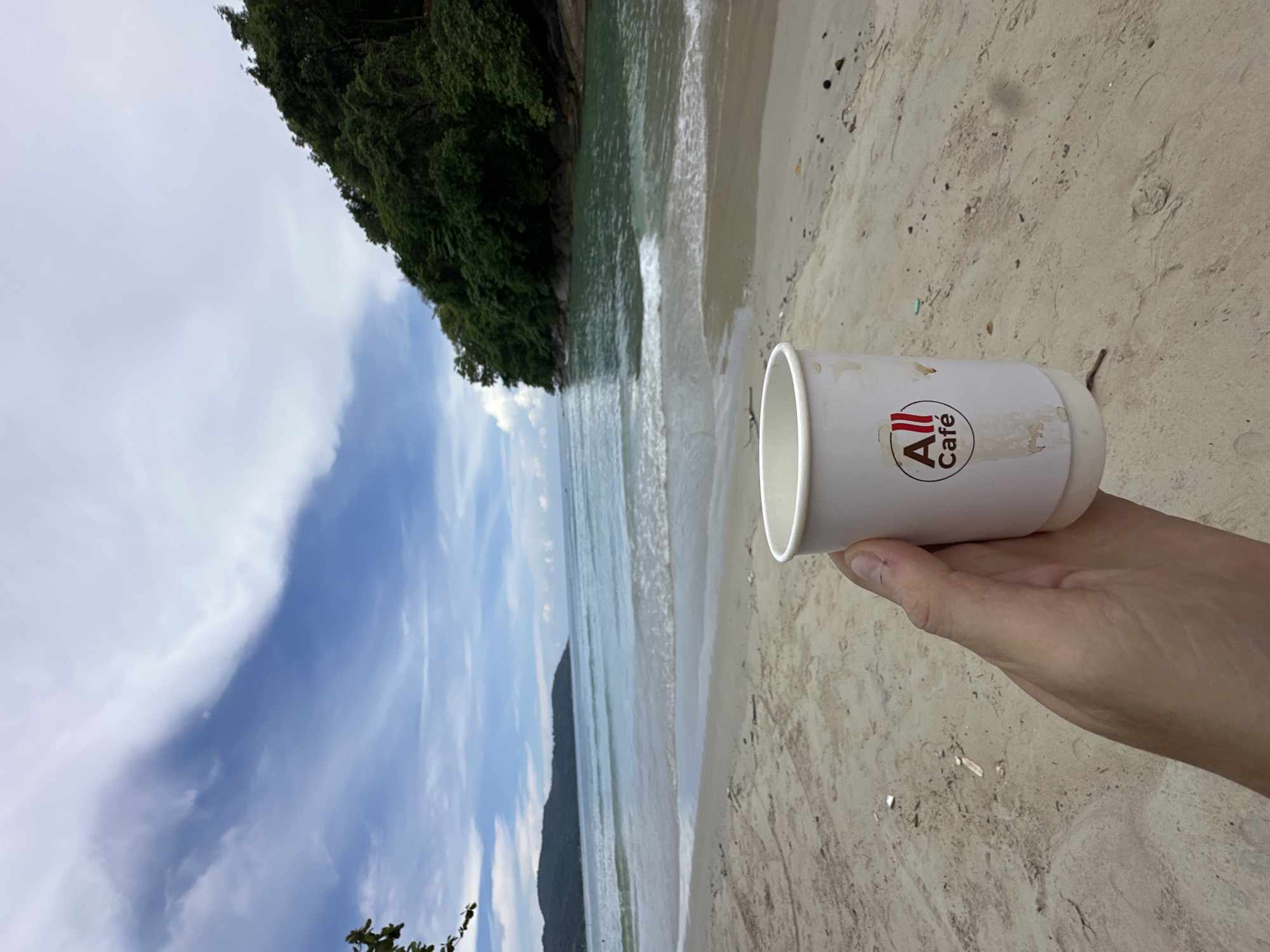 A hand holds a white paper cup with an "All Café" logo on it while standing on a sandy beach in Karon, Thailand. The cup is in the foreground, obscuring some of the sand and its texture. The background shows the ocean with gentle waves, a green, tree-covered hill on the right, and a blue sky with scattered clouds. The water appears calm and turquoise. The beach sand is light brown and shows some debris. The overall scene evokes a relaxed atmosphere, suggesting a break at the beach.