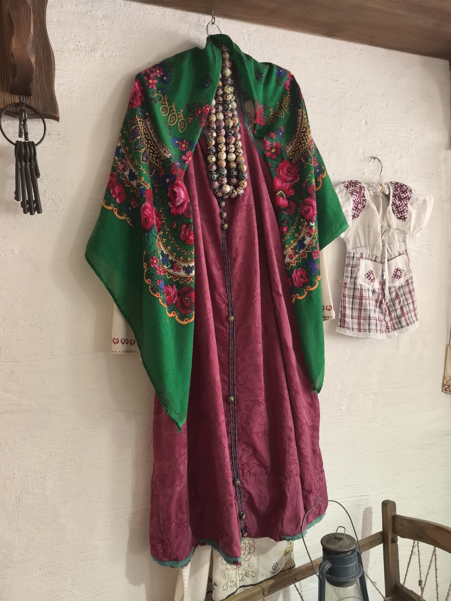 The photograph shows a collection of clothing and decorative items hanging on a white wall, likely indoors. On the left is a set of old keys hanging from a wooden mount. The main focus is a long, maroon or purplish-red garment, possibly a dress or robe, adorned with a necklace of colorful beads and covered with a floral green shawl with flowers and designs on it. To the right of this, hangs a child's dress with a white top and a plaid skirt with embroidered patches. A wooden beam runs along the top of the frame. A lantern is on a piece of furniture in the lower right of the frame. The objects suggest traditional clothing or folk art, possibly on display in a museum or a cultural center. The picture was taken in Russia, in Chaikovsky.