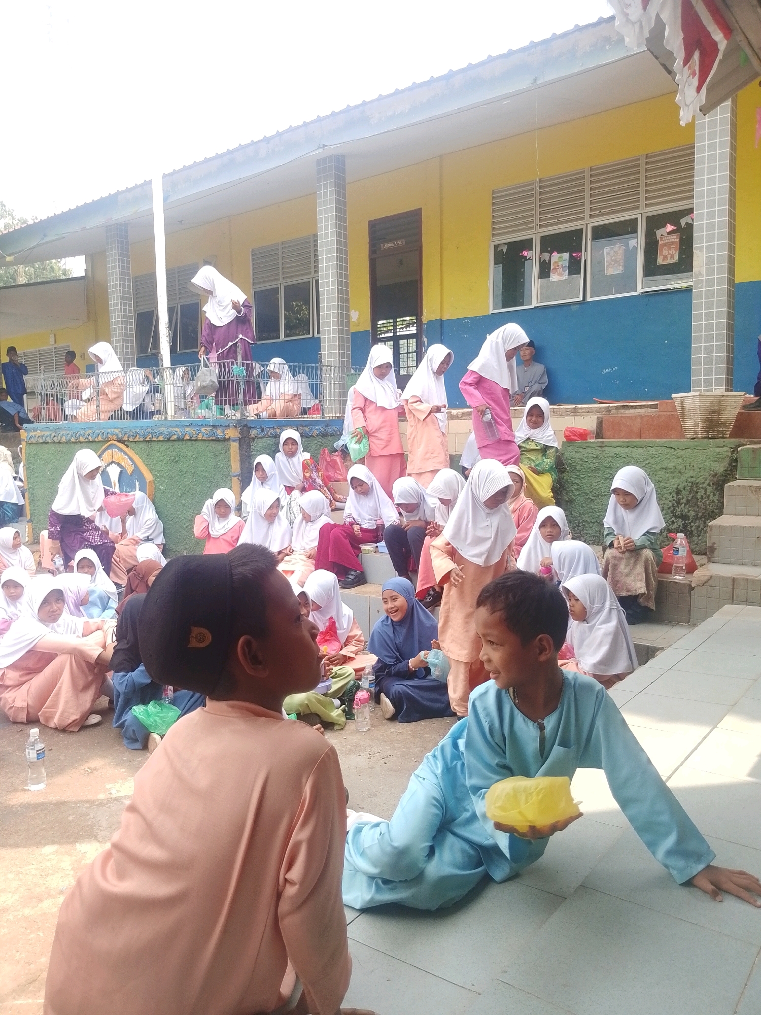 The image depicts a group of children, predominantly girls, gathered outdoors near a school building. The children are mostly wearing headscarves and modest clothing, suggesting a cultural or religious context, possibly a school event or gathering. Many are seated on the ground or on steps, some interacting with each other, while others appear to be listening or observing. The school building in the background is painted yellow and blue, with visible windows and a covered walkway. The overall environment appears to be a schoolyard or an outdoor educational space in Batam, Indonesia. The presence of water bottles and some plastic bags suggests a casual event.