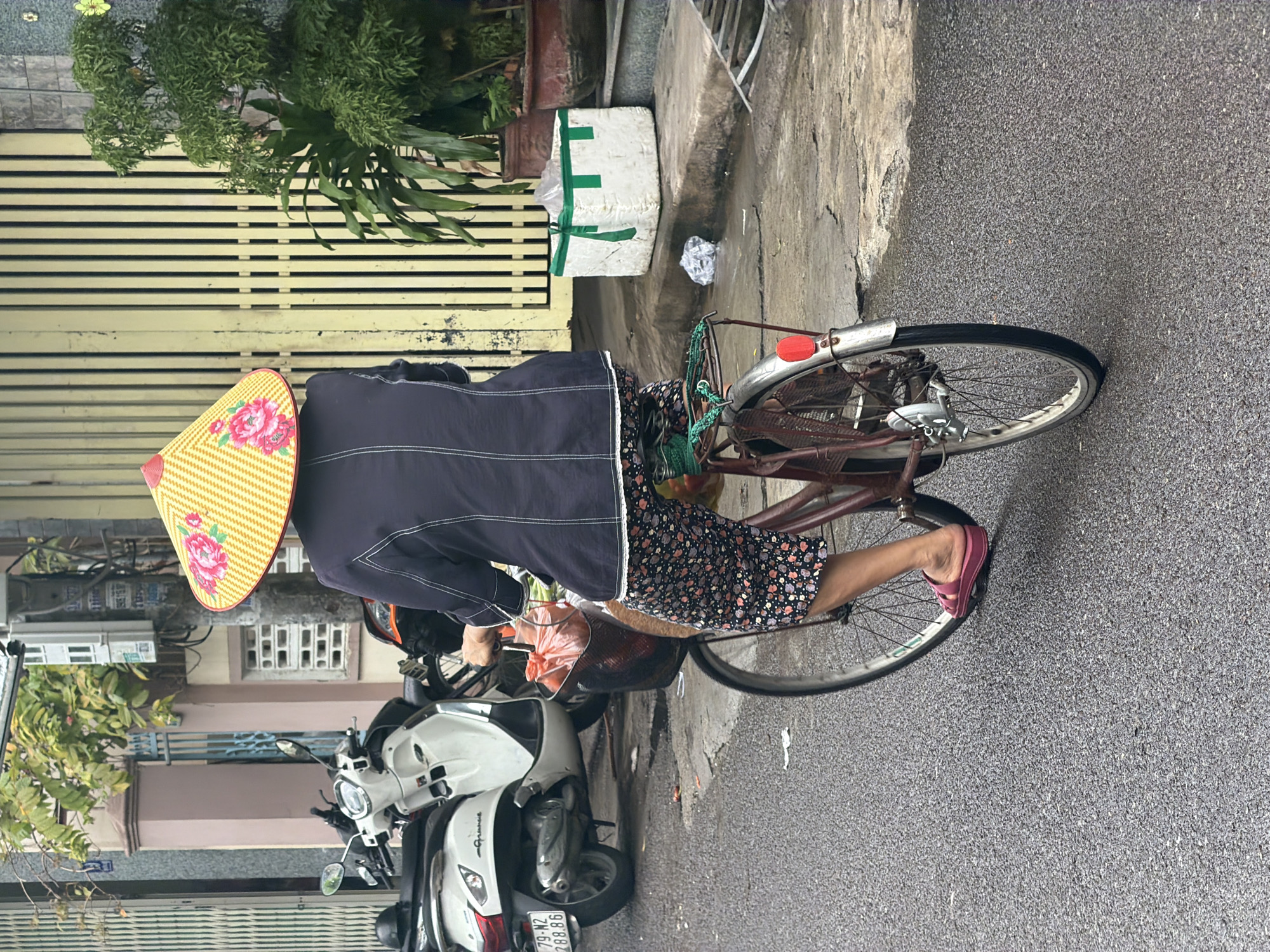 The image shows an outdoor urban street scene in Nha Trang Ward, Vietnam, centered on a person on a bicycle. The individual, seen from behind and slightly to the left, wears a traditional Vietnamese conical hat (nón lá) featuring a vibrant yellow color and a large red/pink floral design. They are dressed in a dark blue long-sleeved top with visible white stitching and a patterned floral skirt. Pink sandals are on their feet, with one foot touching the damp asphalt road, suggesting the cyclist is either stopped or moving slowly.
The bicycle has a brown frame and silver fenders; its front basket contains a red plastic bag and other items. To the left, a building with light-colored horizontal slat gates is visible, adorned with green potted plants. A white rectangular object secured with green tape rests on the concrete curb. Further left and behind the cyclist, several motorbikes are present, including a white scooter with the brand name "Grande" and a visible license plate "79-N2 288.86". The street surface is dark, damp asphalt. The scene depicts a commonplace moment of daily life in a Vietnamese urban setting during what appears to be daytime, possibly under overcast skies or after rain.