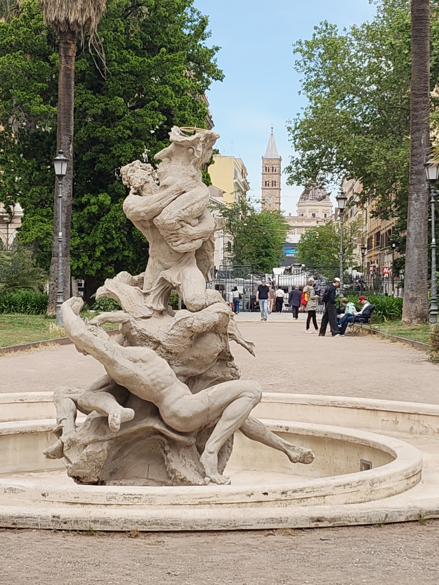 The image depicts a large stone fountain sculpture in a park setting. The sculpture features human figures in a dynamic, intertwined composition. In the background, a pedestrian walkway lined with trees and lampposts is visible. People are walking along the path, and some are sitting on benches. Buildings are seen in the distance, including a tall, narrow tower with a clock and a domed structure. This scene is located in Rome, Italy.