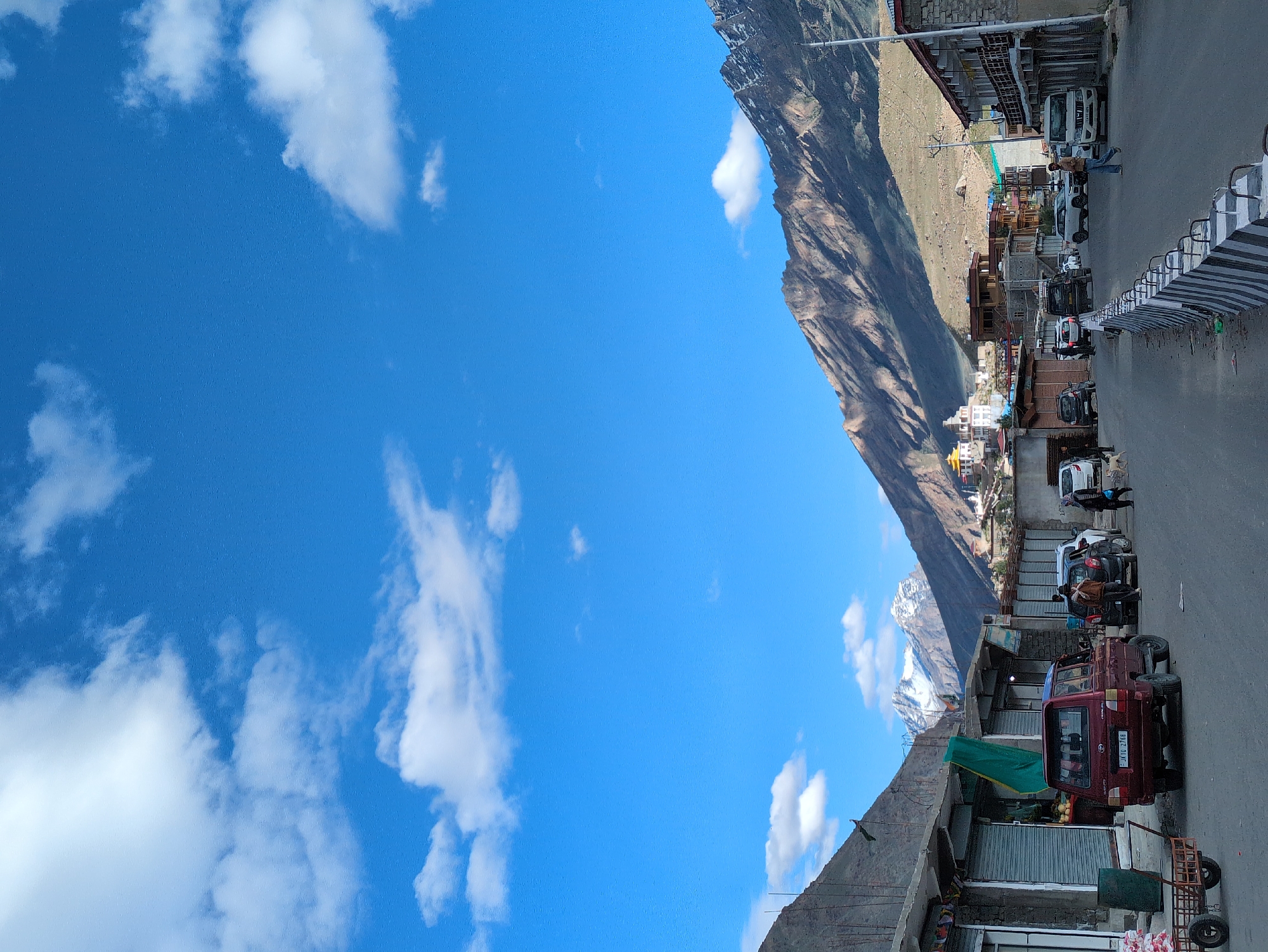 A wide-angle, full shot captures a street scene in Padum, India, beneath a vibrant blue sky dotted with white clouds. The street runs through a valley with steep, rugged mountains on either side. The mountainsides are largely barren, with some patches of scrubby vegetation. The town of Padum is visible nestled in the distance, characterized by white and yellow buildings, some with distinctive architectural features, situated on the slopes of the mountains.
In the foreground, the asphalt street is lined with shops and buildings. The shops on the left have their metal shutters down, while the buildings on the right appear to be multi-story structures with balconies. A small red SUV with license plate "JK10 2768" is parked on the left side of the street. Several other vehicles, including cars and what appears to be a white SUV, are also present on the street, suggesting moderate activity.
A few individuals are walking along the street. On the right side, a person wearing a brown jacket and blue jeans is walking away from the camera. Further down the street, another person is walking towards the camera. On the left side, near the shops, a few more figures are visible, some appearing to be near the parked red SUV. A dog is also present on the street near the cars.
To the left of the red SUV, a rustic wooden cart with wheels is parked, laden with white sacks. The street is demarcated by a concrete barrier with alternating black and white stripes, serving as a protective measure. The overall impression is a bustling, yet serene, mountain town environment under a clear, sunny sky.