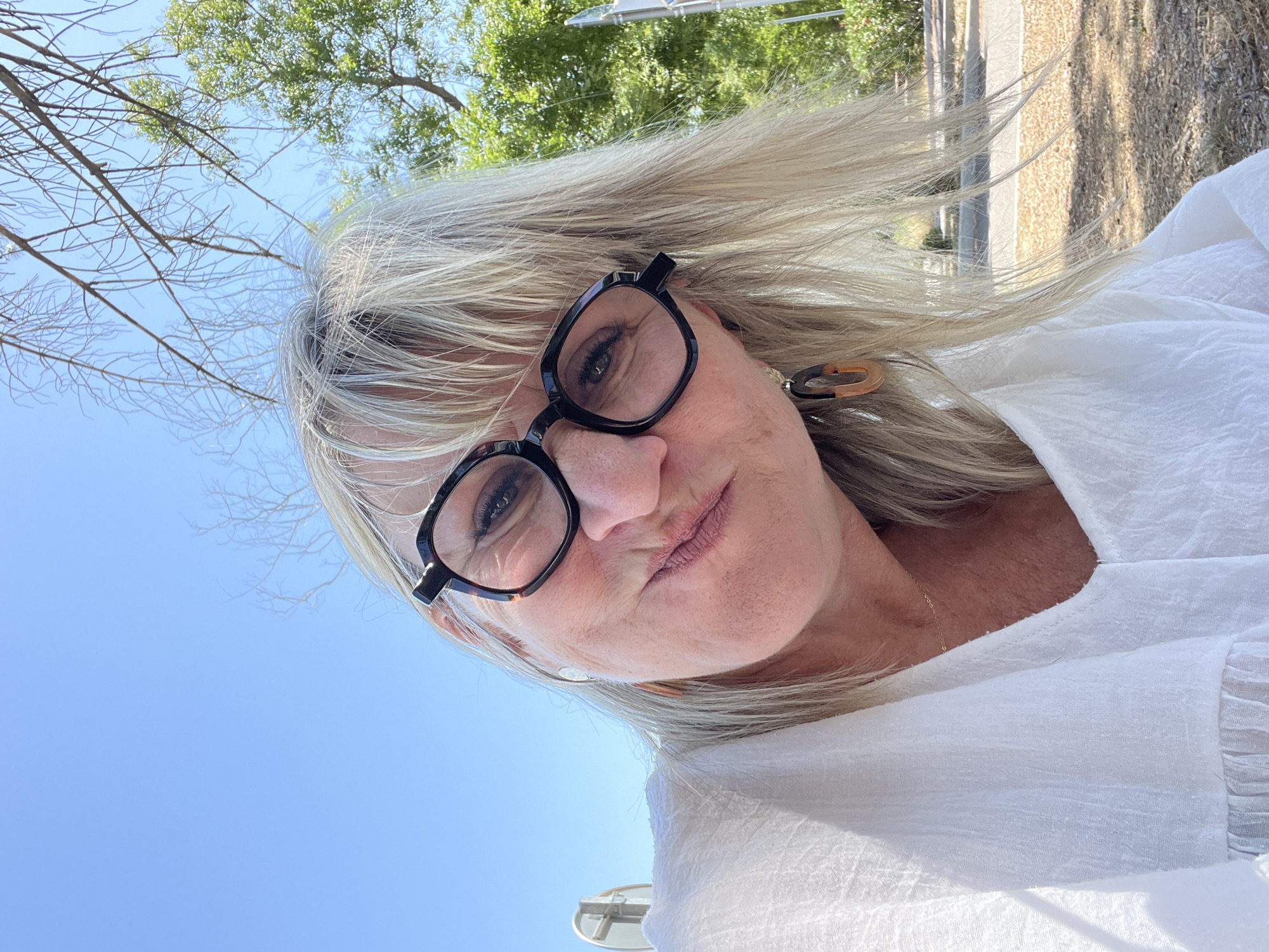 A woman with blonde hair and glasses takes a selfie outdoors. She is wearing a white top and her hair is blowing in the wind. The background shows a bright blue sky, green trees, and a building with textured walls. The sunlight suggests it is daytime, likely a sunny day. The woman has a slightly pursed lips expression, and her eyes are looking towards the camera. She is wearing a pair of black-rimmed glasses and a tortoiseshell-patterned earring. A thin gold chain necklace is visible around her neck. The setting appears to be a natural or semi-outdoor environment, possibly a garden or courtyard.
