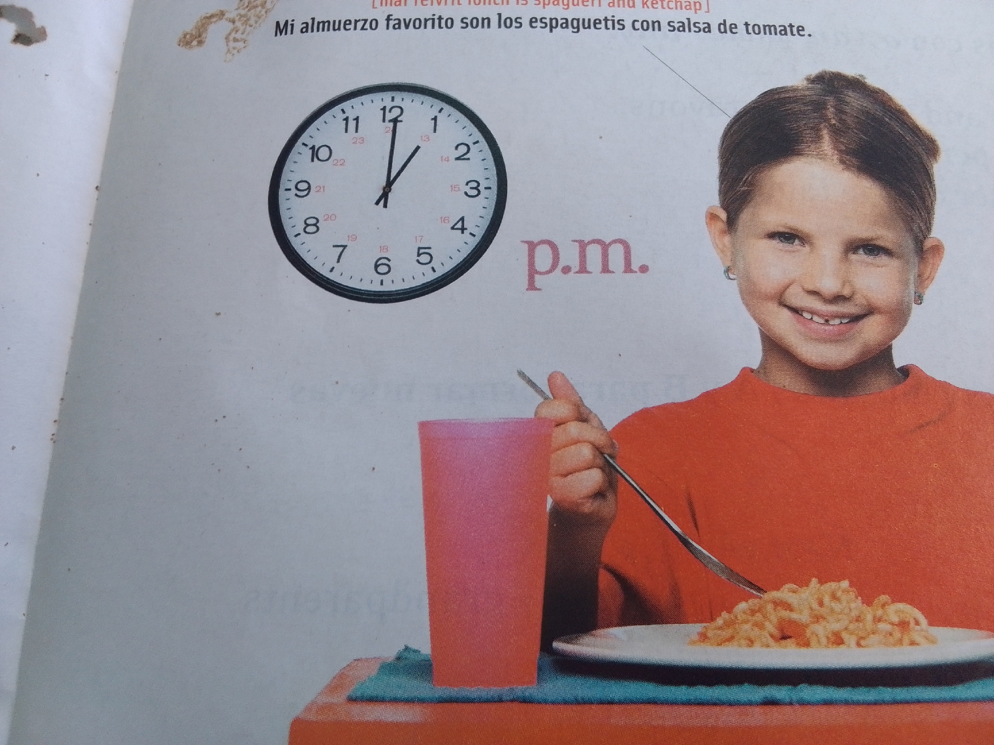 The image shows a young girl with a wide smile, missing a front tooth, wearing a bright orange shirt and small hoop earrings. She is looking directly at the camera. In front of her is a plate of pasta, presumably spaghetti, with a fork poised to take a bite. To her left, a tall, pink plastic cup sits on a blue and orange placemat. A clock on the left side of the image shows the time as approximately 12:03. Text in Spanish above the image translates to "My favorite lunch is spaghetti with tomato sauce." There is no information in the image that places the scene in La Tagua, Venezuela.
